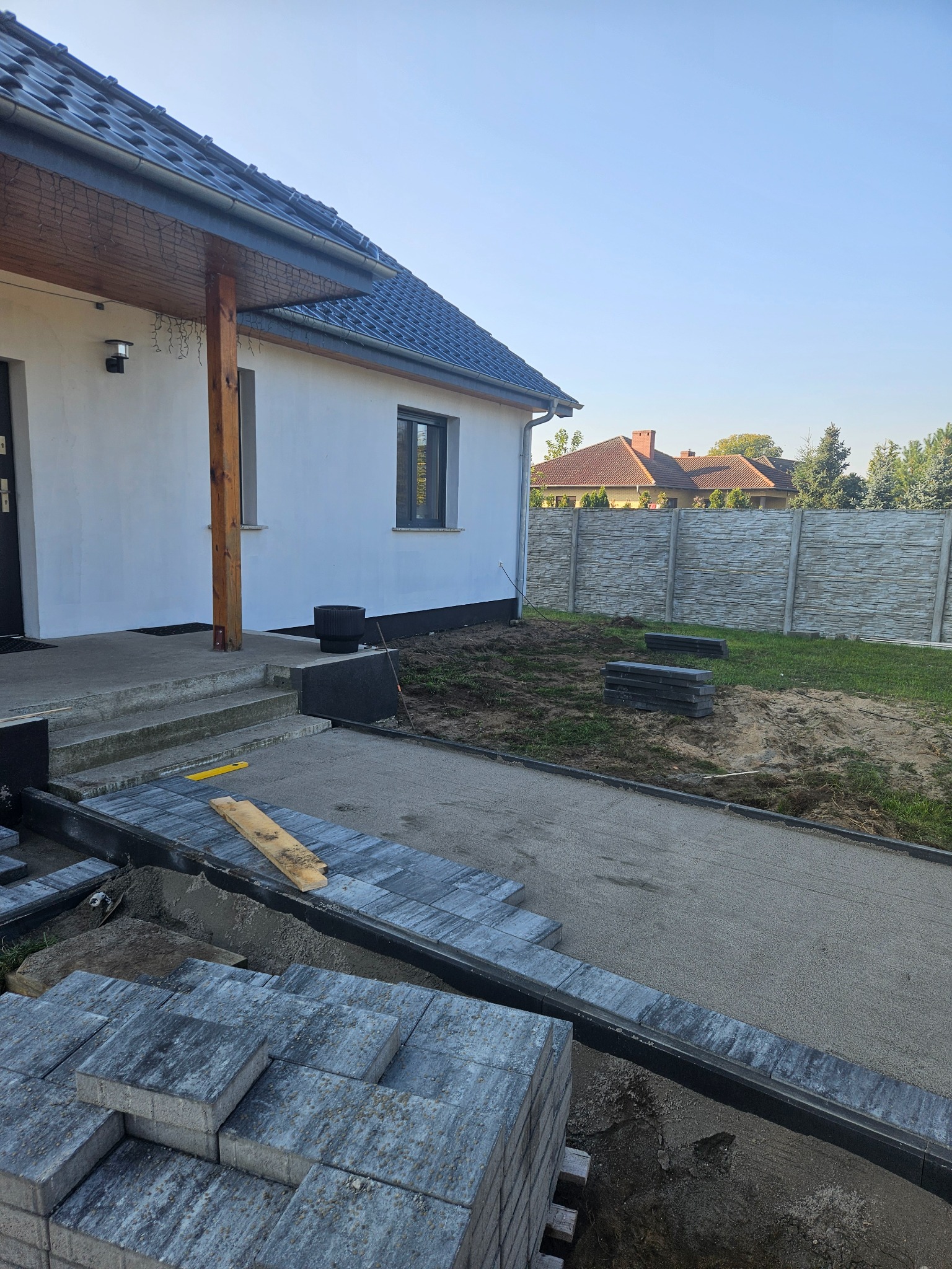 Ułożona część ścieżki z szarej kostki brukowej na posesji z białym domem i betonowym ogrodzeniem w tle. Kostka czeka na ułożenie. Widoczny poziomica.