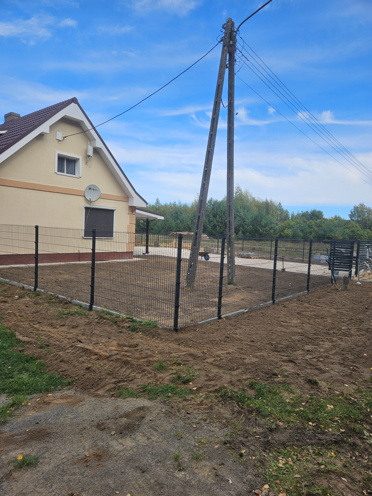 Nowoczesne ogrodzenie panelowe z podmurówką betonową, otaczające posesję z domem jednorodzinnym. Widoczne słupy i siatka w kolorze grafitowym, świeżo wyrównana ziemia wewnątrz ogrodzenia.