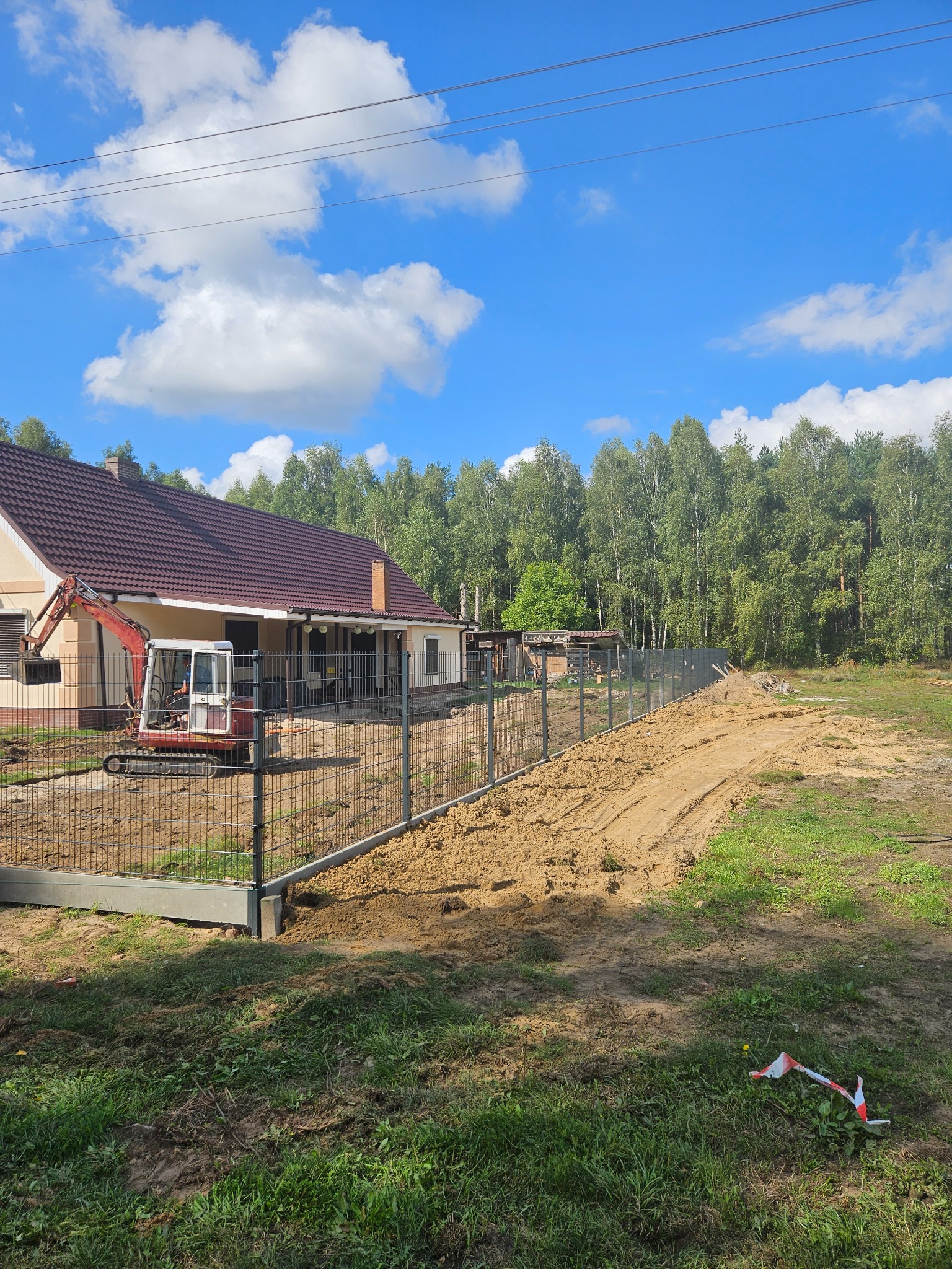 Nowoczesne ogrodzenie panelowe z podmurówką na nierównym terenie, z widocznym mini-koparką i domem w tle, w słoneczny dzień. Widoczne ślady prac ziemnych.