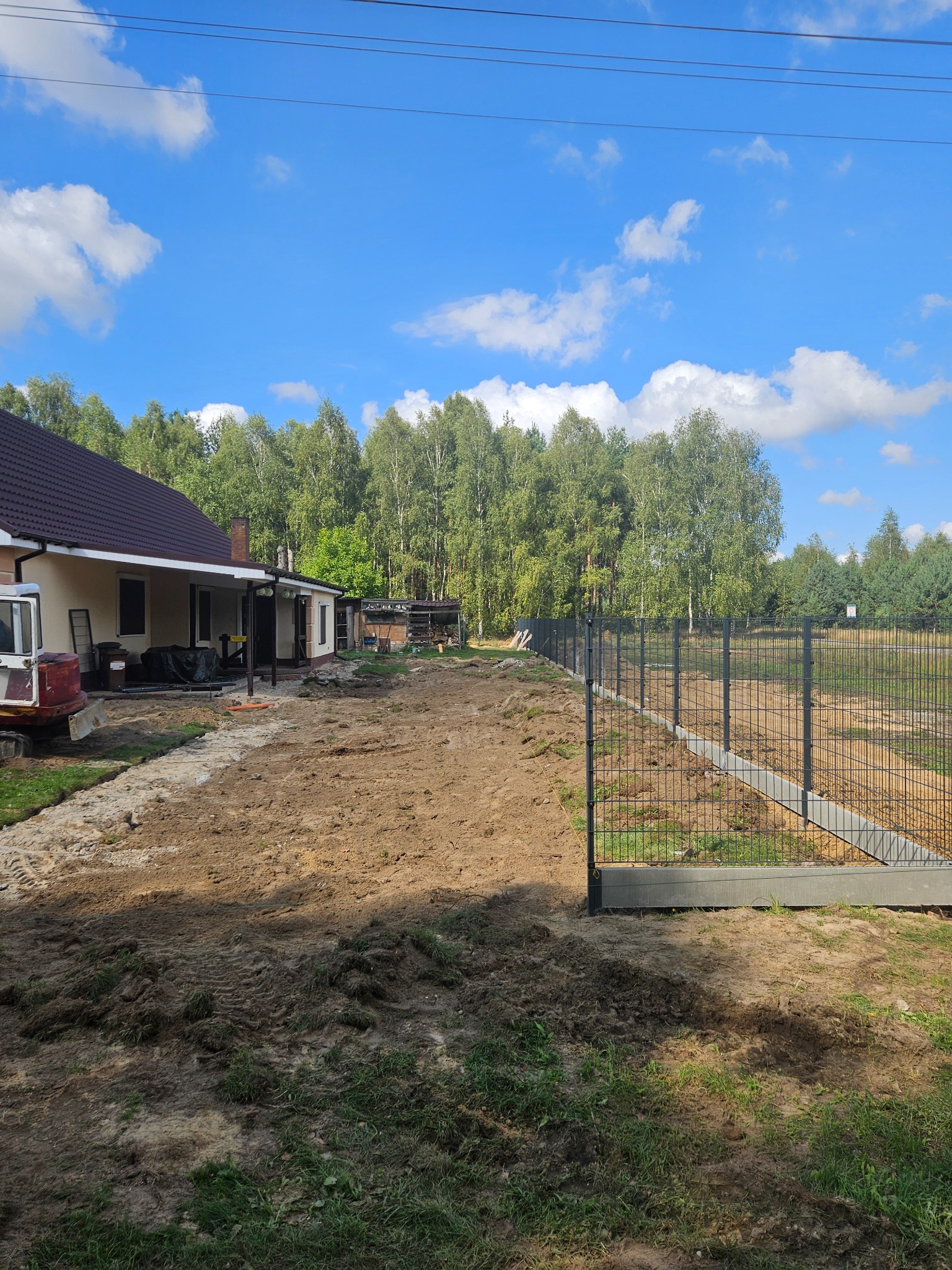 Montaż ogrodzenia panelowego na betonowej podmurówce, widok na posesję z domem i przygotowanym terenem pod trawnik. Słoneczny dzień, błękitne niebo.