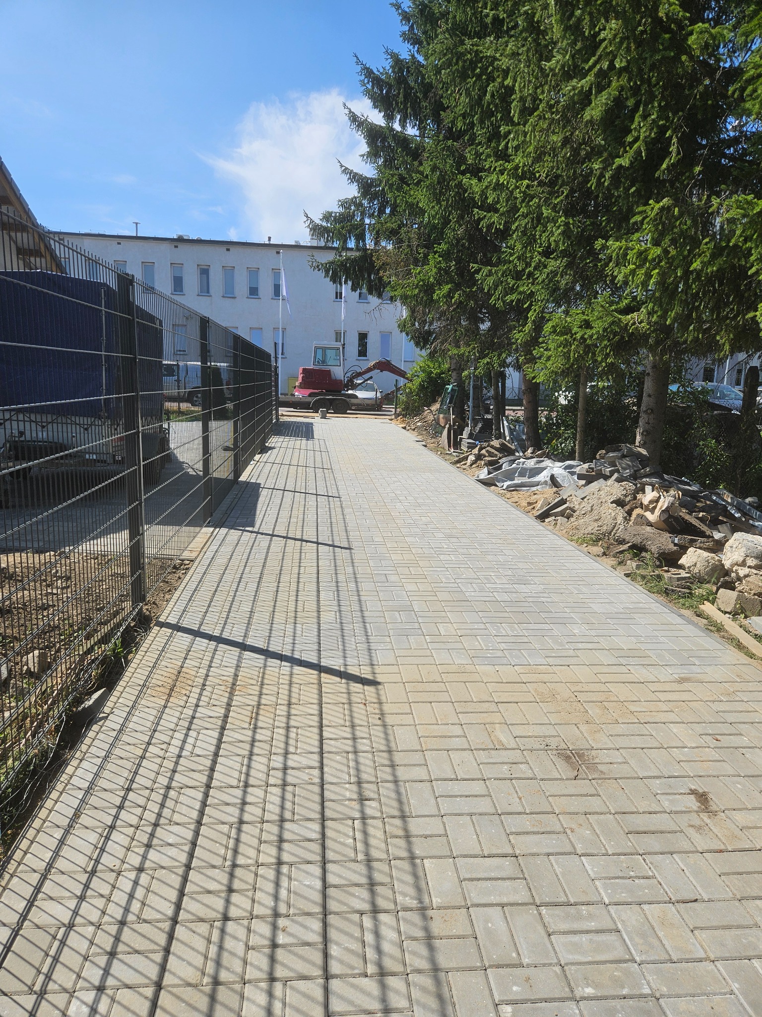 Ułożona kostka brukowa wzdłuż ogrodzenia, z widocznym w tle małym koparką i budynkiem. Cień ogrodzenia tworzy wzór na kostce. Praca w toku.