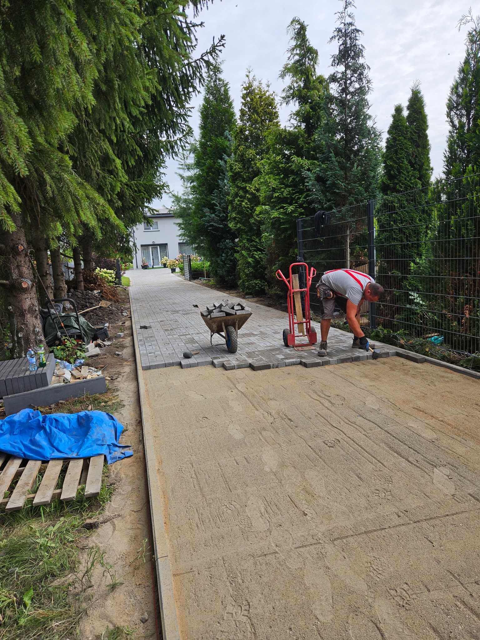 Pracownik w trakcie układania szarej kostki brukowej na podsypce piaskowej. Widoczny wózek z kostką, narzędzia i fragment posesji z zielenią i ogrodzeniem.