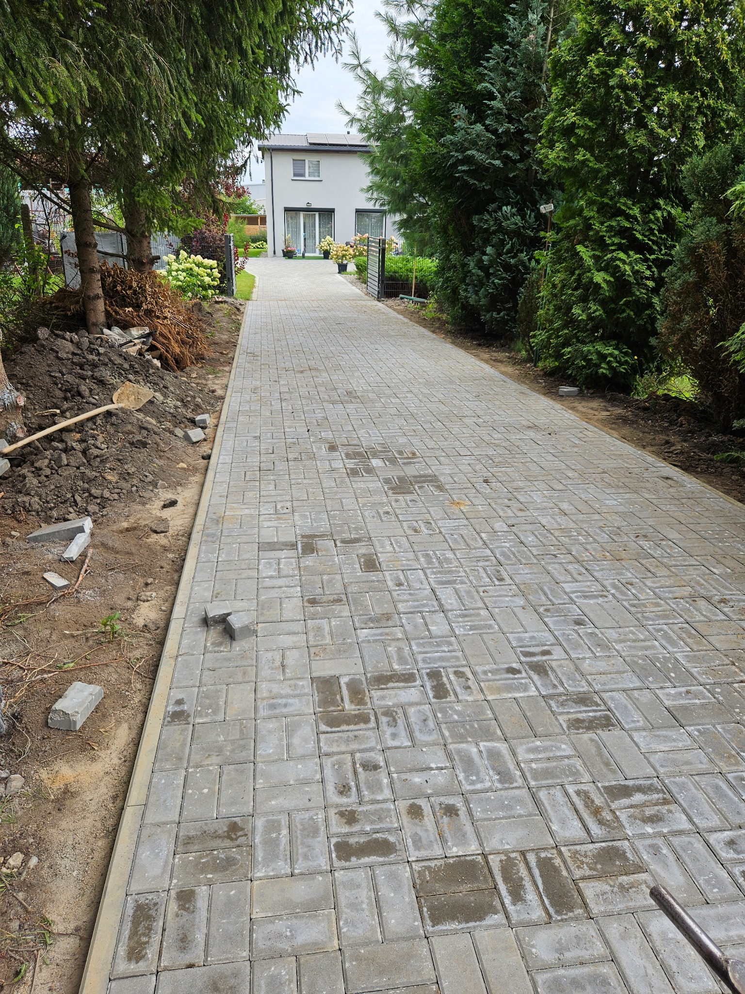 Długa, szara ścieżka z kostki brukowej prowadzi do domu. Po lewej stronie widoczna ziemia z narzędziami, po prawej krzewy. Ujęcie z perspektywy.