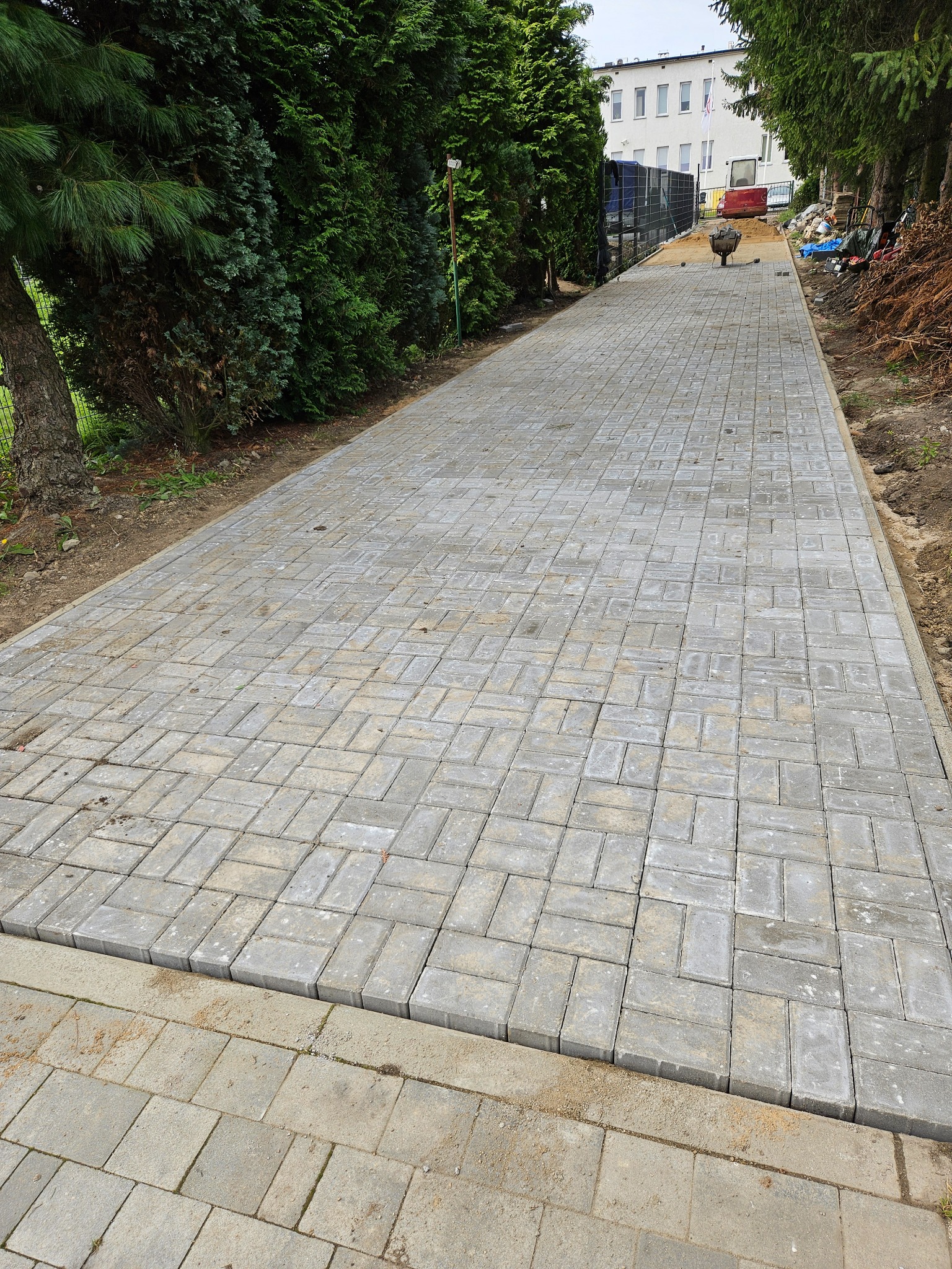 Ułożona, szara kostka brukowa tworząca ścieżkę wzdłuż zielonych drzew i budynku w tle. Widoczny fragment terenu przygotowanego pod dalsze prace.