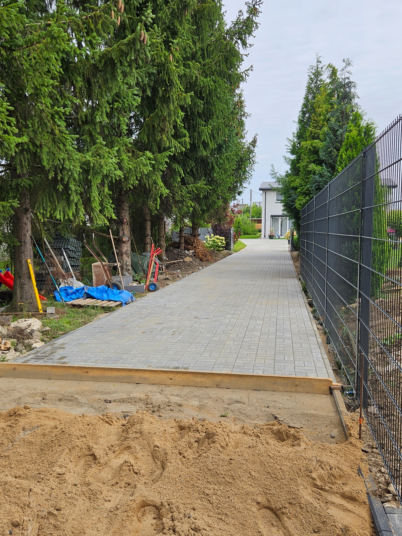 Ścieżka z szarej kostki brukowej w trakcie budowy, widoczny piasek i drewniane obramowanie, w tle zielone drzewa i czarne ogrodzenie panelowe.