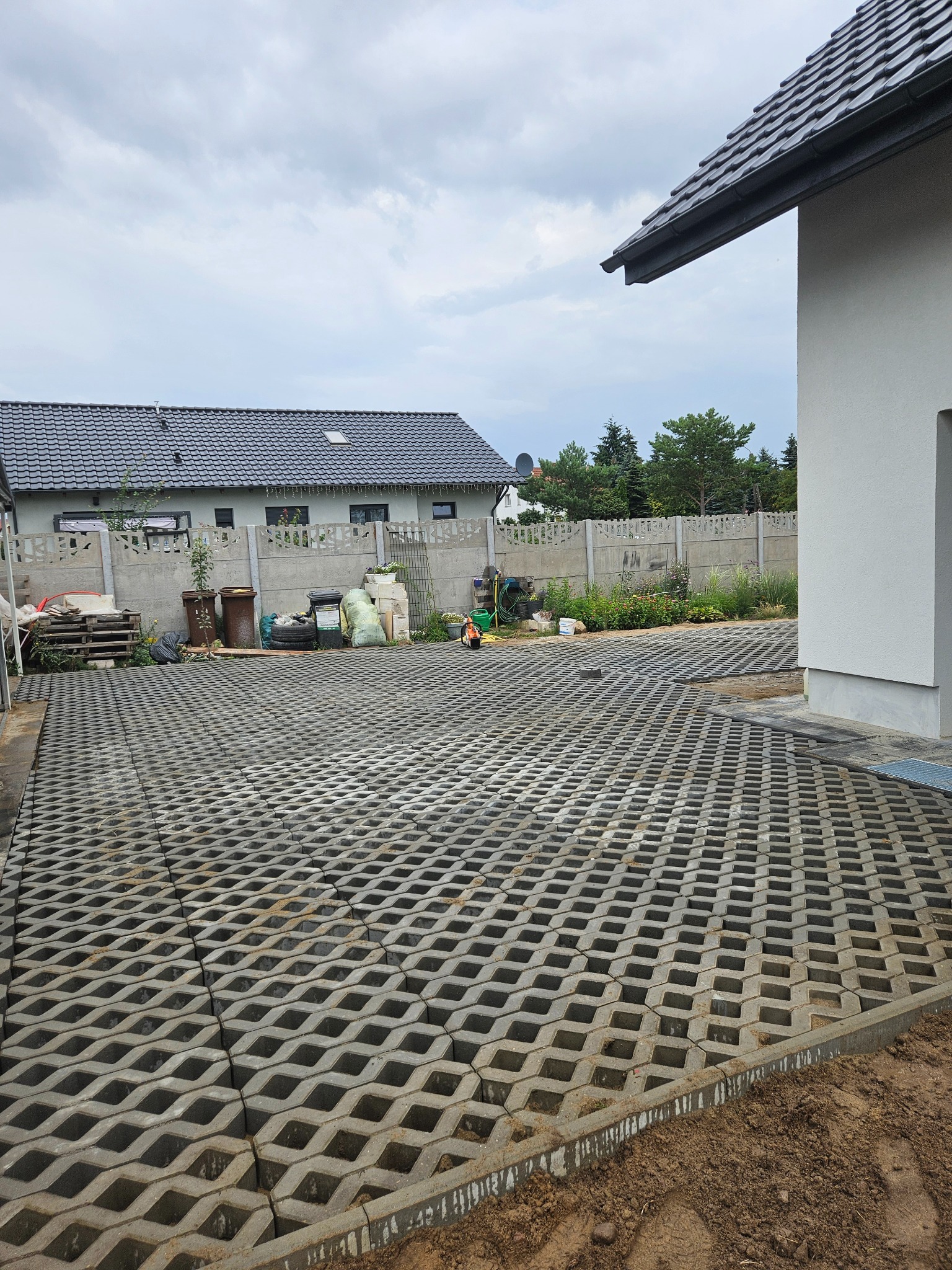 Ułożona szara ażurowa kostka brukowa na podjeździe, widok z góry. W tle dom z szarą dachówką i betonowe ogrodzenie. Częściowo widoczny narożnik budynku.