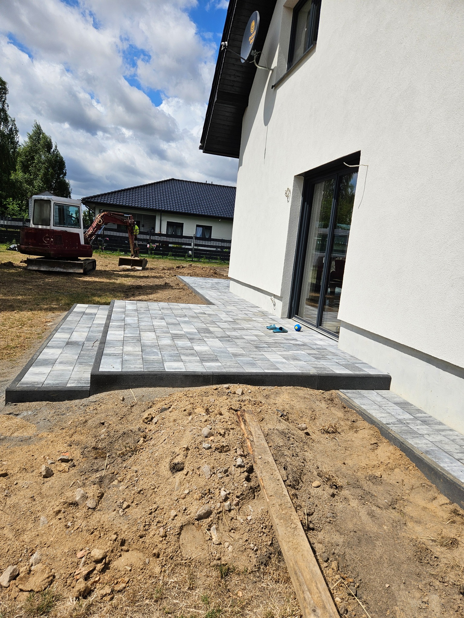 Ułożona kostka brukowa na tarasie domu jednorodzinnego, z widocznym mini-koparką w tle. Częściowo wykopana ziemia w pierwszym planie.