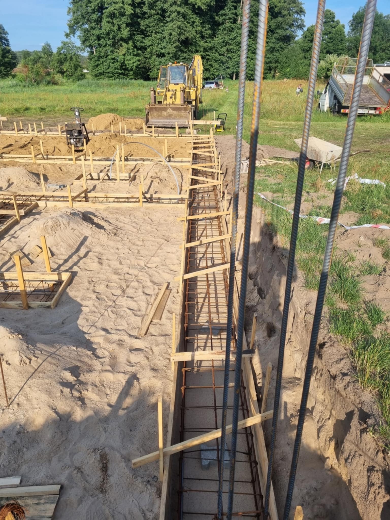 Fundamenty domu w trakcie budowy: szalunki, zbrojenie, wykop. W tle koparka i ubijarka. Widok z góry na plac budowy w słoneczny dzień.