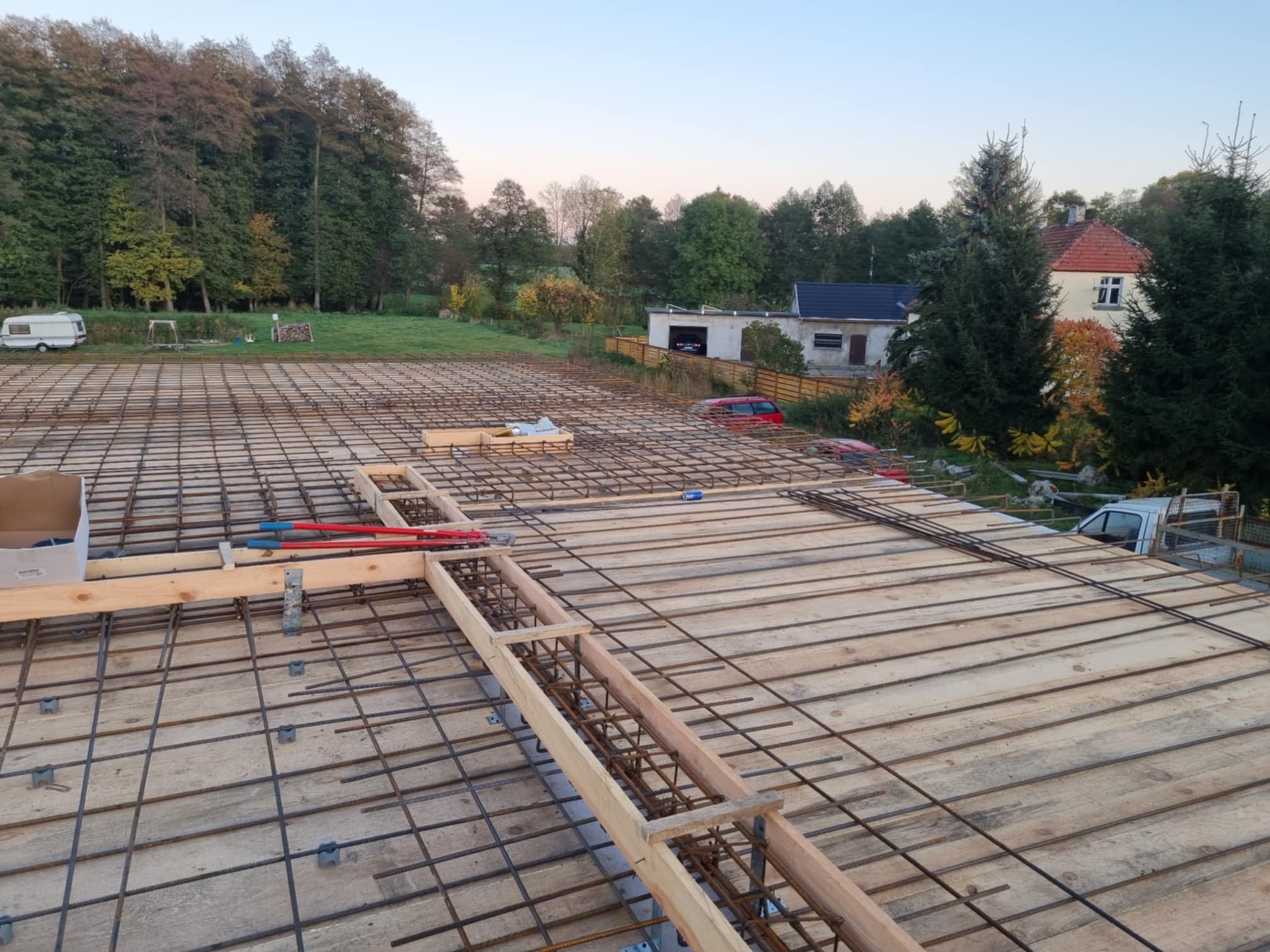 Zbrojenie stropu domu jednorodzinnego z widocznymi elementami szalunku i siatką z prętów stalowych. W tle zabudowa jednorodzinna i zieleń.