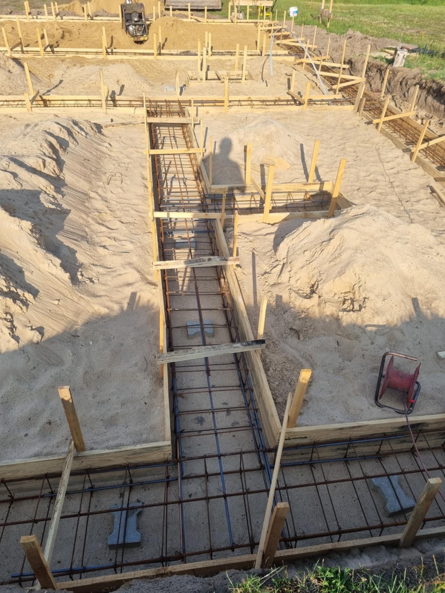 Zbrojenie fundamentów domu jednorodzinnego w wykopie, drewniane szalunki, piasek, widoczny cień osoby, zwijarka kabla w tle. Etap budowy.