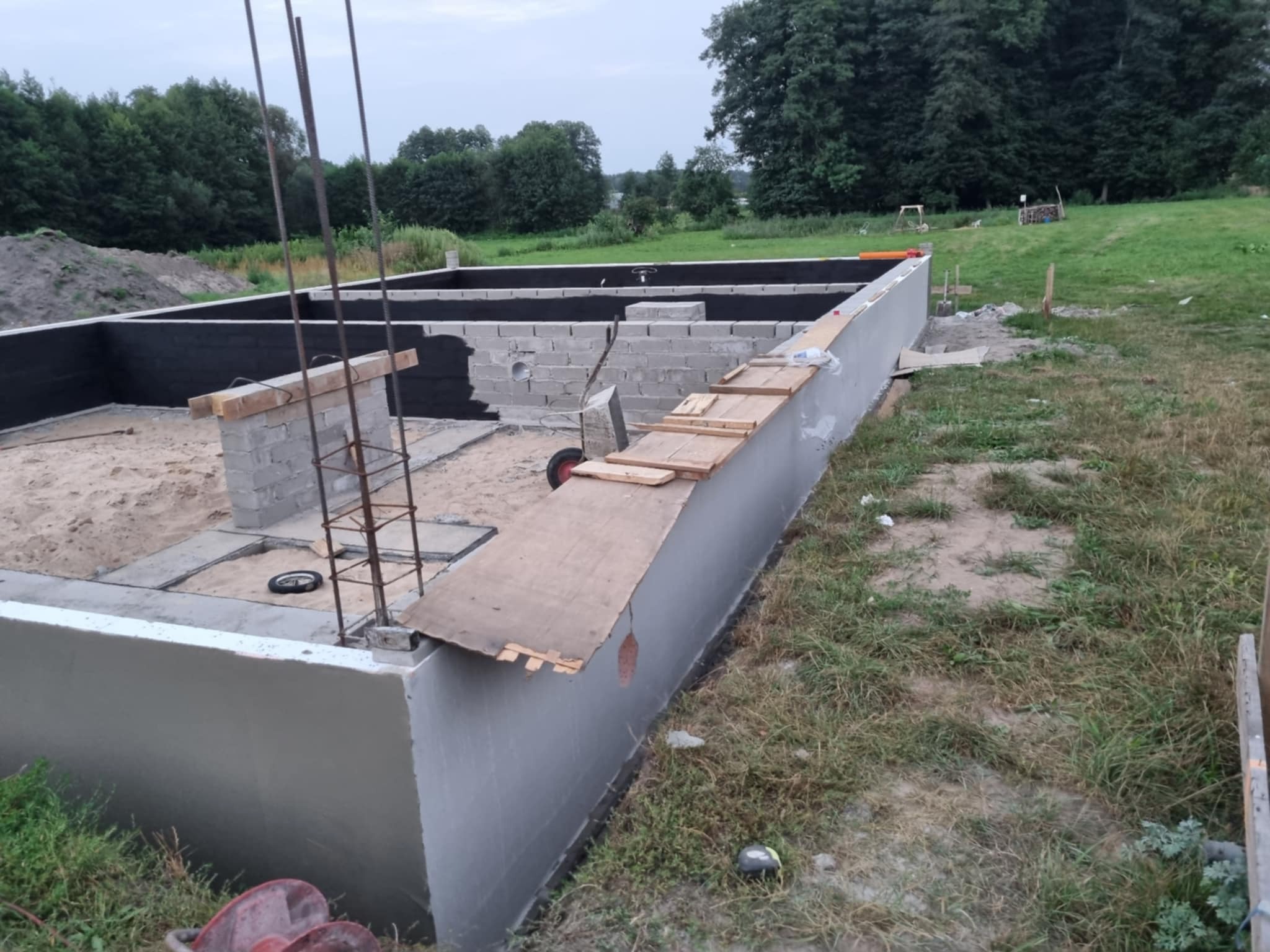 Fundamenty domu w trakcie budowy: betonowe ściany, izolacja, zbrojenie. Widoczne bloczki betonowe i piasek wewnątrz. Tło: trawa i drzewa.