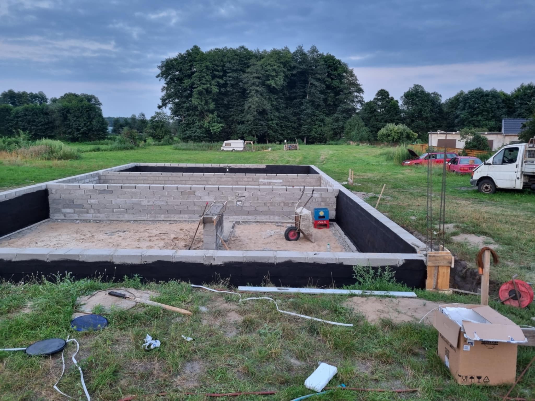 Fundamenty domu w trakcie budowy, zabezpieczone czarną izolacją, na tle zieleni i pochmurnego nieba. Widoczne narzędzia i materiały budowlane.