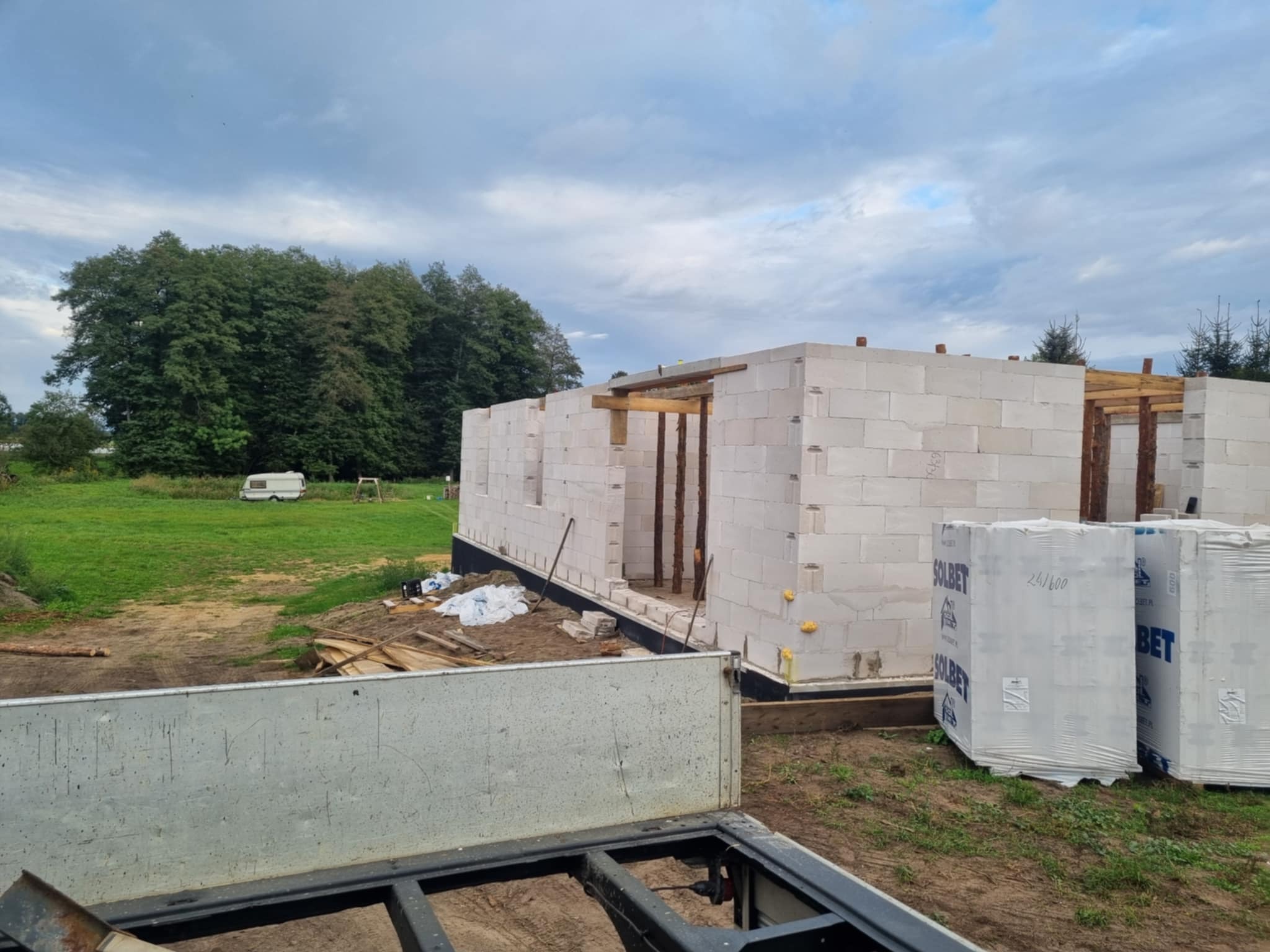 Wznoszenie ścian parterowego domu z białych bloczków Solbet. Widoczne pakiety bloczków, fragment przyczepy i zielone otoczenie. Budowa w toku.