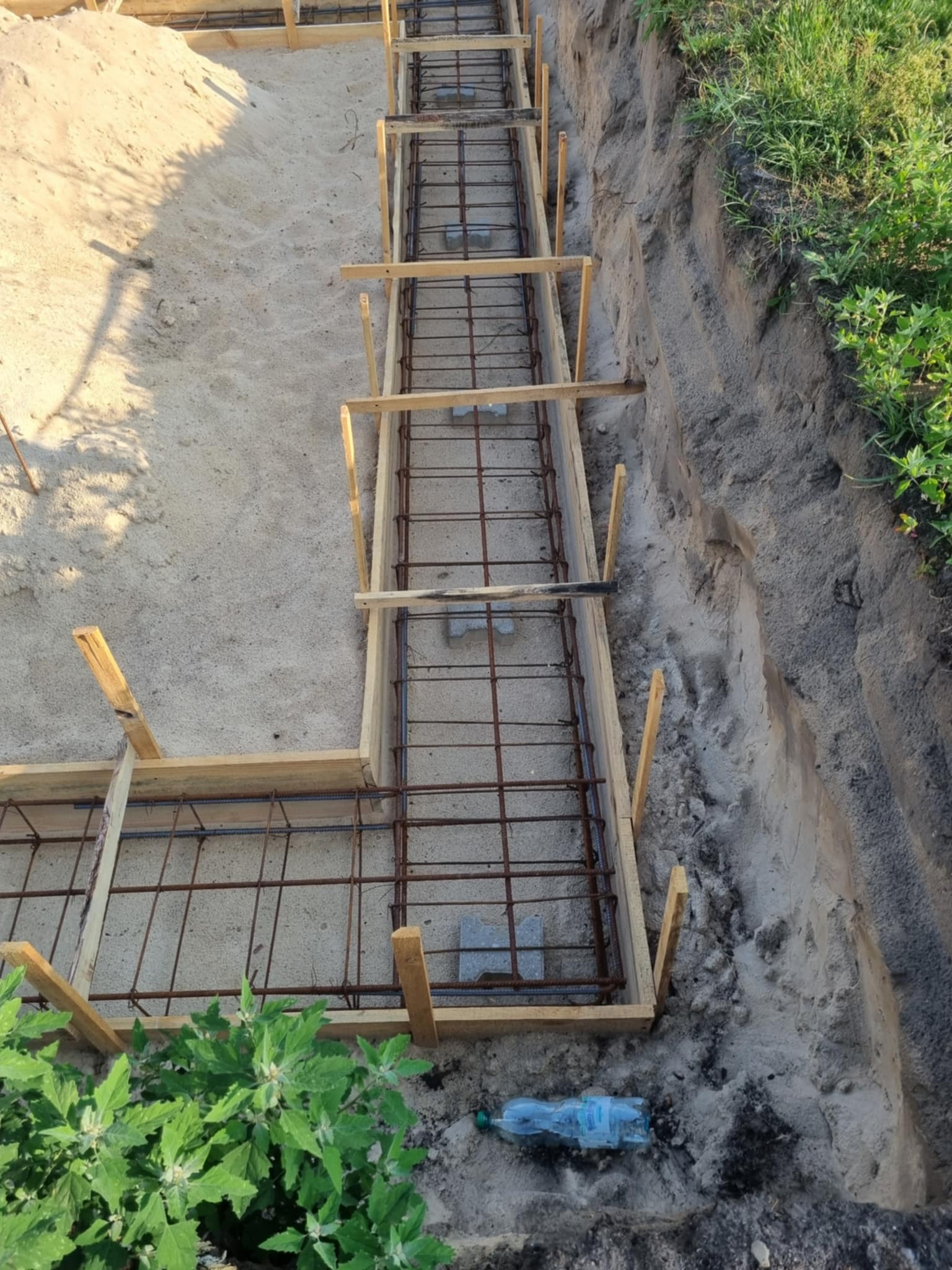 Zbrojenie fundamentów w wykopie, drewniane szalunki, widoczne pręty zbrojeniowe i betonowe podkładki dystansowe. W tle ziemia i roślinność.