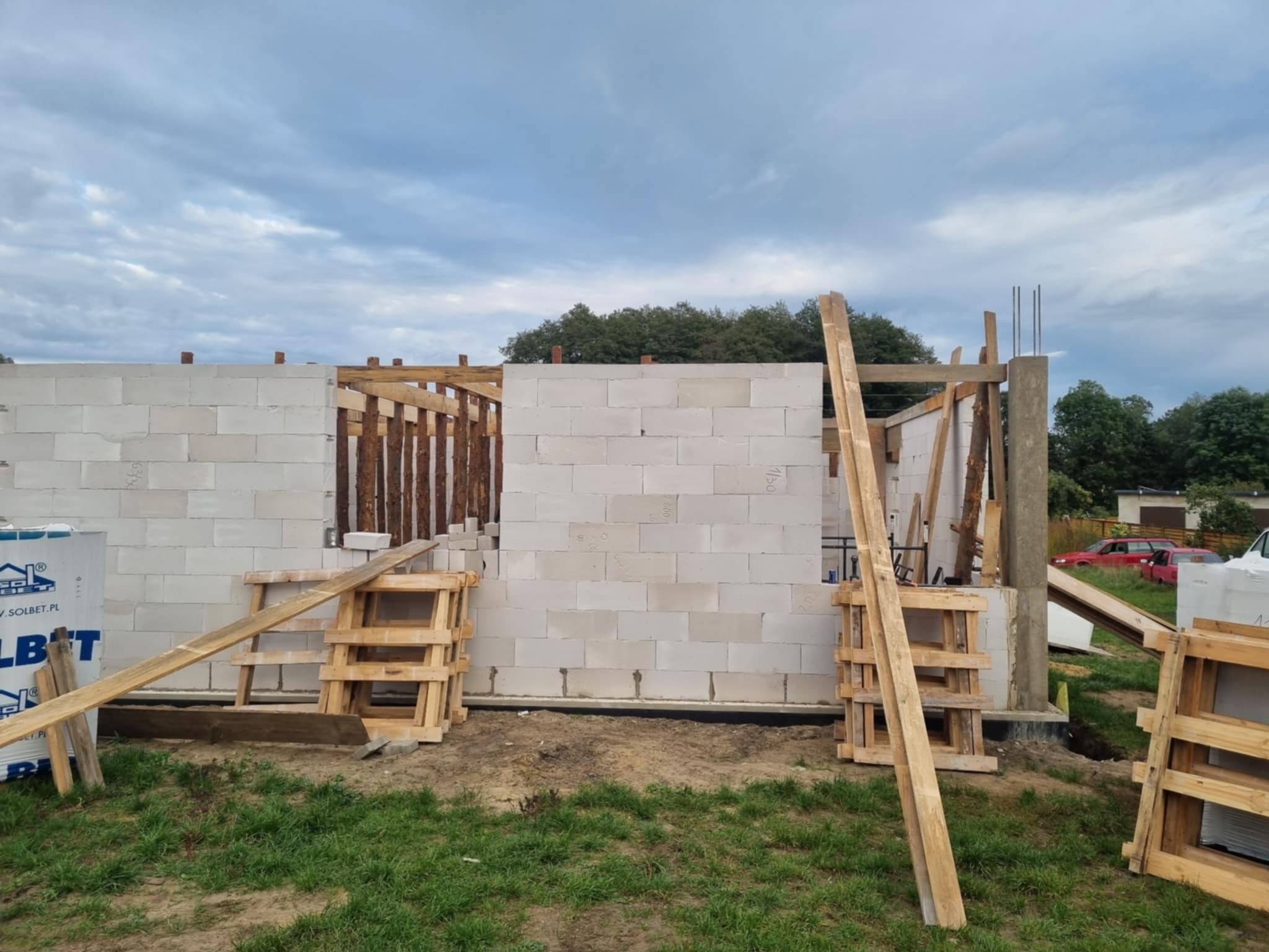 Budowa domu z białych bloczków: ściany w trakcie wznoszenia, drewniane elementy konstrukcyjne, palety budowlane na trawiastym terenie. Widok na niebo i drzewa w tle.