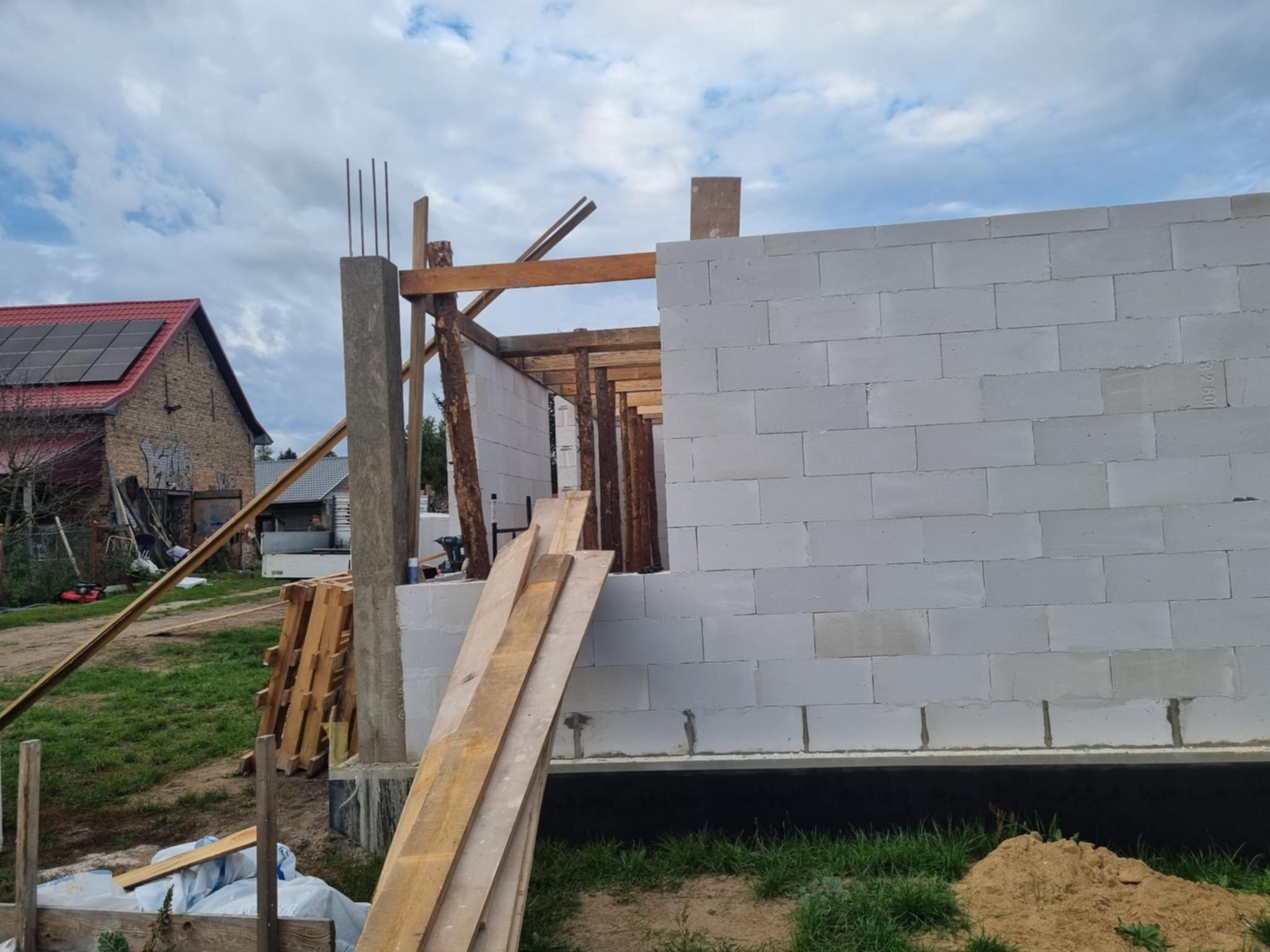 Wznoszenie ścian domu z bloczków betonu komórkowego, widoczne drewniane elementy konstrukcyjne i zbrojenie. W tle budynek z panelami słonecznymi na dachu.