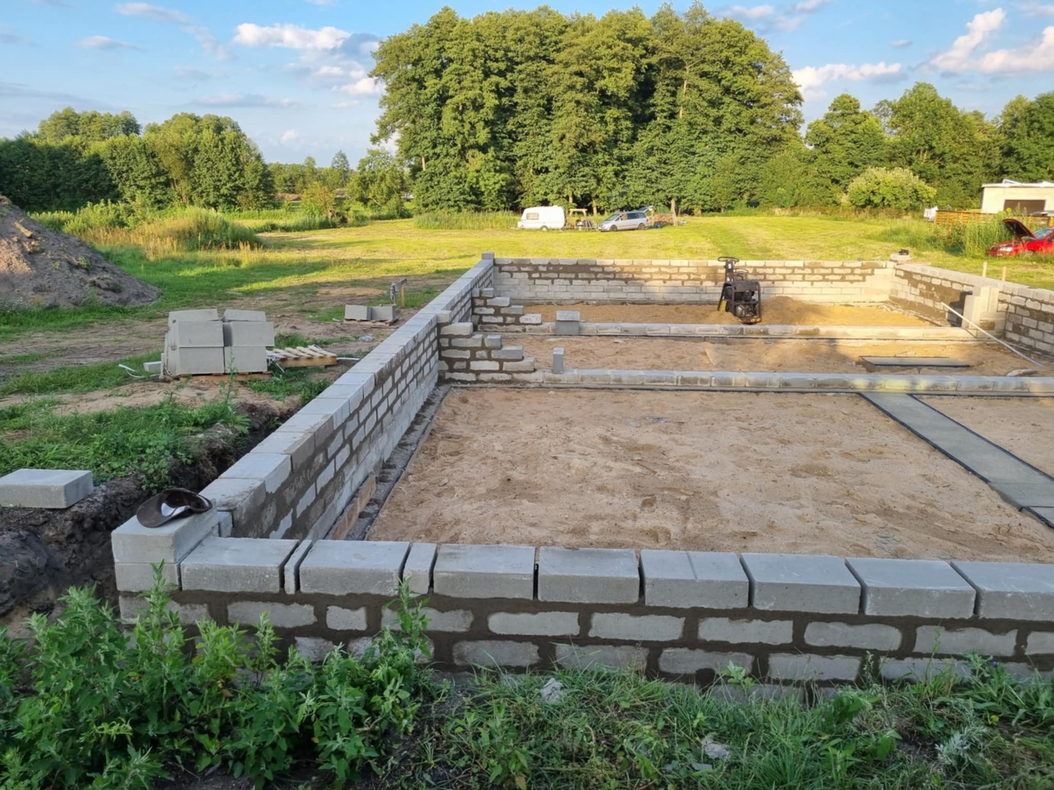 Budowa fundamentów domu z bloczków betonowych na piaszczystym podłożu. Widoczny ubijak wibracyjny i zapas bloczków. Tło: zieleń drzew i błękitne niebo.