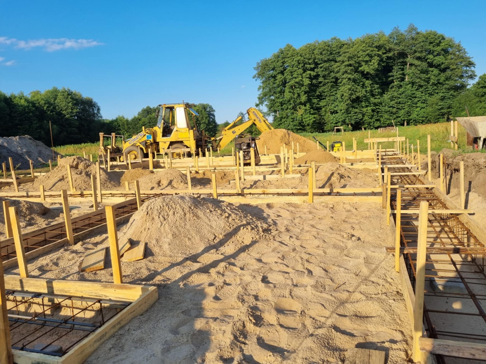 Fundamenty domu w trakcie budowy z widocznym zbrojeniem, drewnianym szalunkiem i koparkami w tle, na tle zieleni i błękitnego nieba.