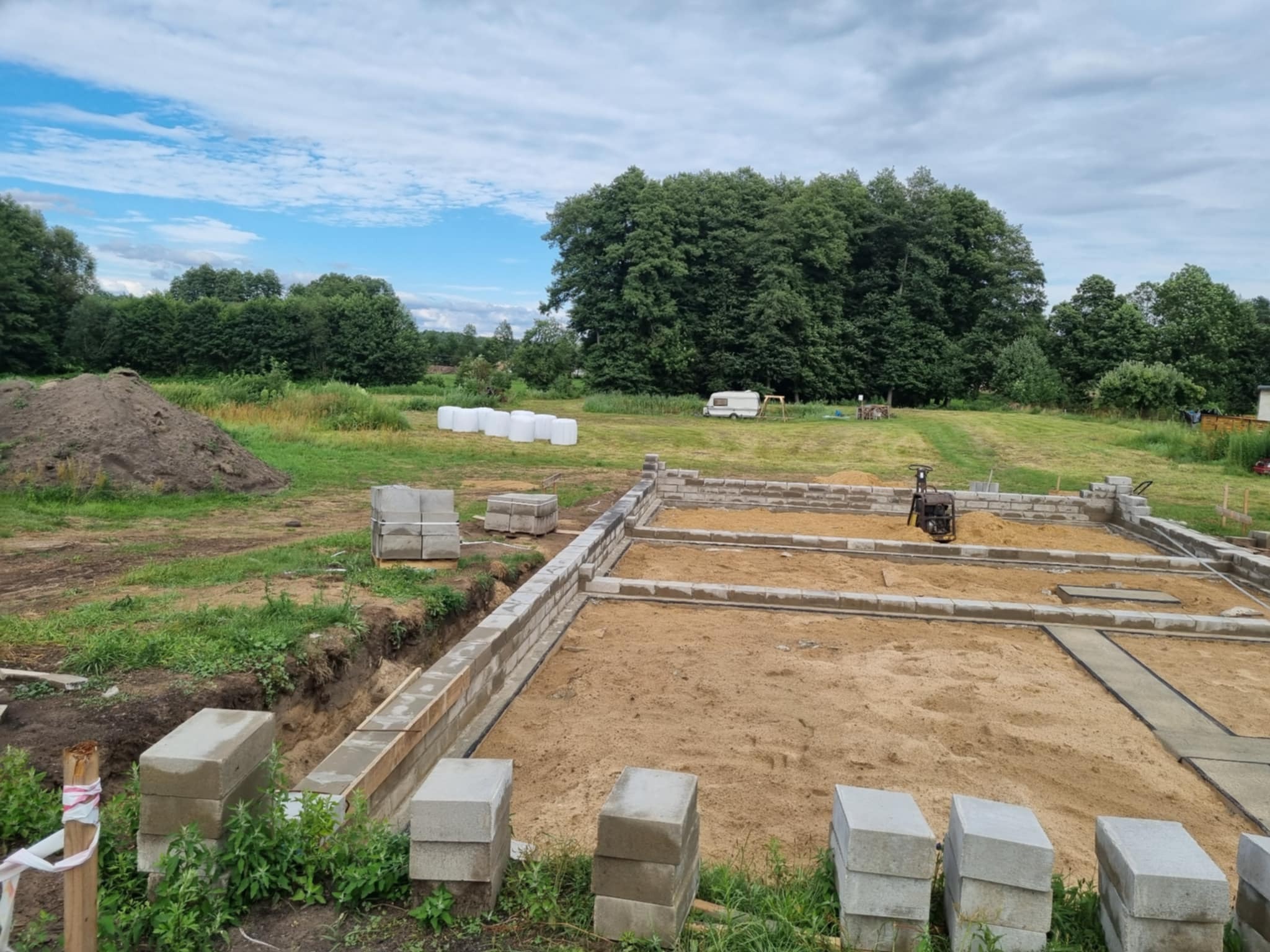 Fundamenty domu w trakcie budowy, wykonane z szarych bloczków betonowych, na piaszczystym podłożu. W tle zielone drzewa i błękitne niebo z chmurami.