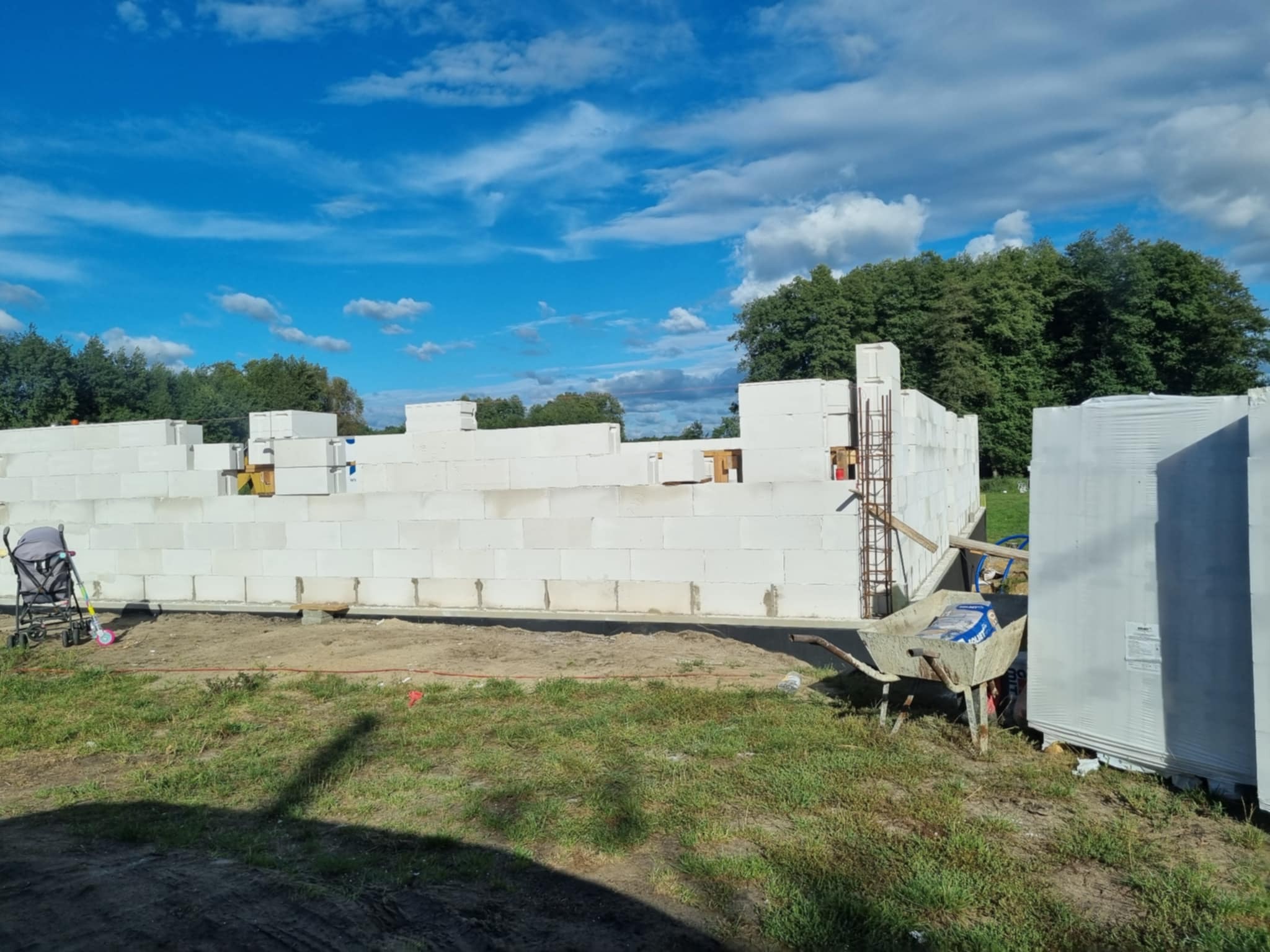 Wznoszenie ścian domu z bloczków betonu komórkowego Ytong. Widoczne zbrojenia, taczka i materiały budowlane na działce z zieloną trawą i błękitnym niebem.