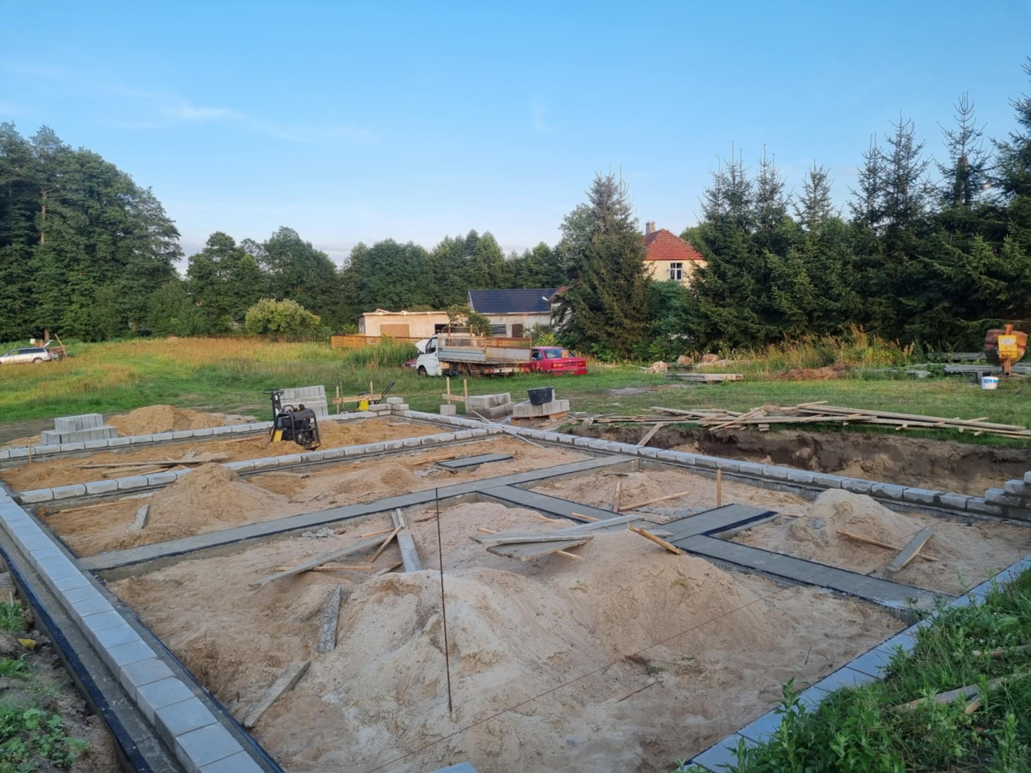 Fundamenty domu w trakcie budowy w Gorzowie Wielkopolskim. Widoczne bloczki fundamentowe, piasek i częściowo zalane ławy. W tle zieleń i zabudowania.