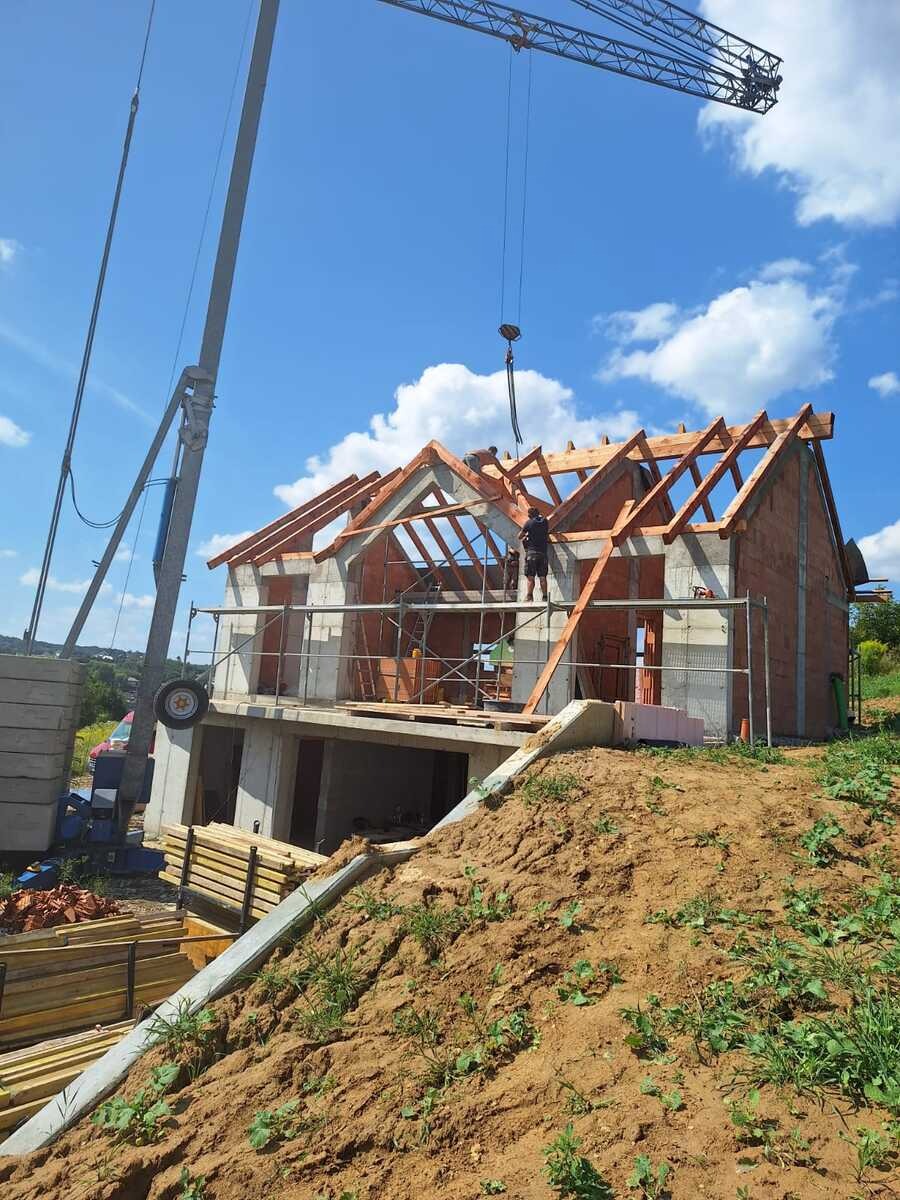 Budowa domu murowanego z czerwonej cegły. Widoczny dźwig, rusztowania i pracownicy montujący drewnianą konstrukcję dachu na tle błękitnego nieba.