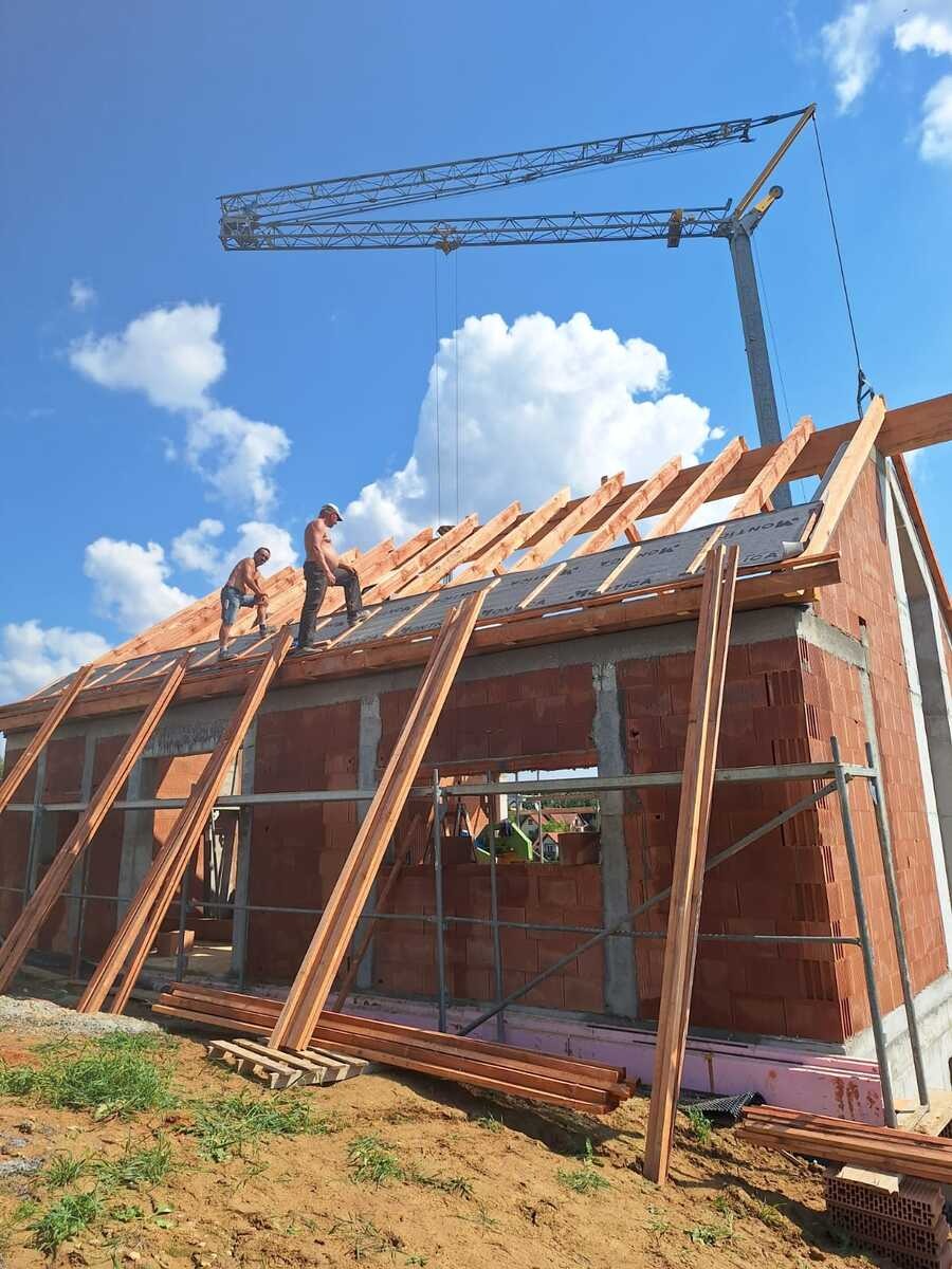 Budowa domu z czerwonej cegły: murarze montują drewnianą konstrukcję dachu na tle błękitnego nieba z chmurami i żurawia budowlanego.