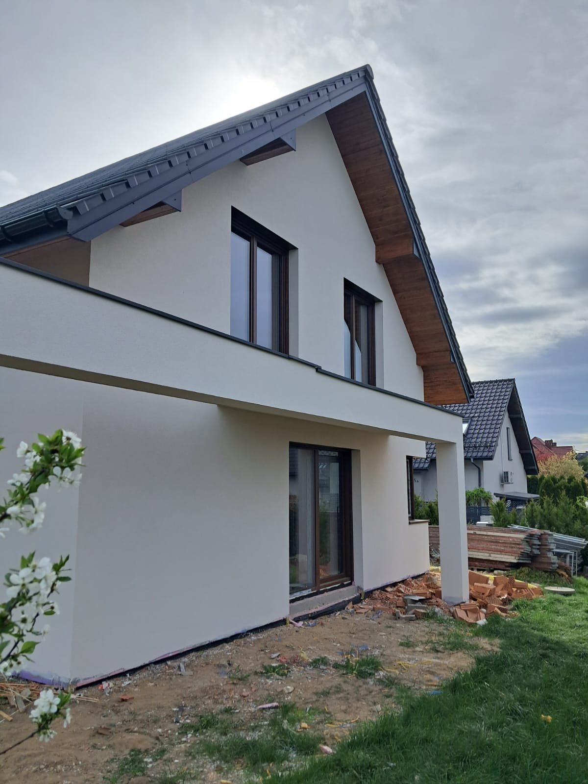 Nowoczesny dom jednorodzinny z jasną elewacją i ciemną dachówką, widok z boku, z fragmentem kwitnącej gałęzi na pierwszym planie. Częściowo zagospodarowany teren wokół budynku.