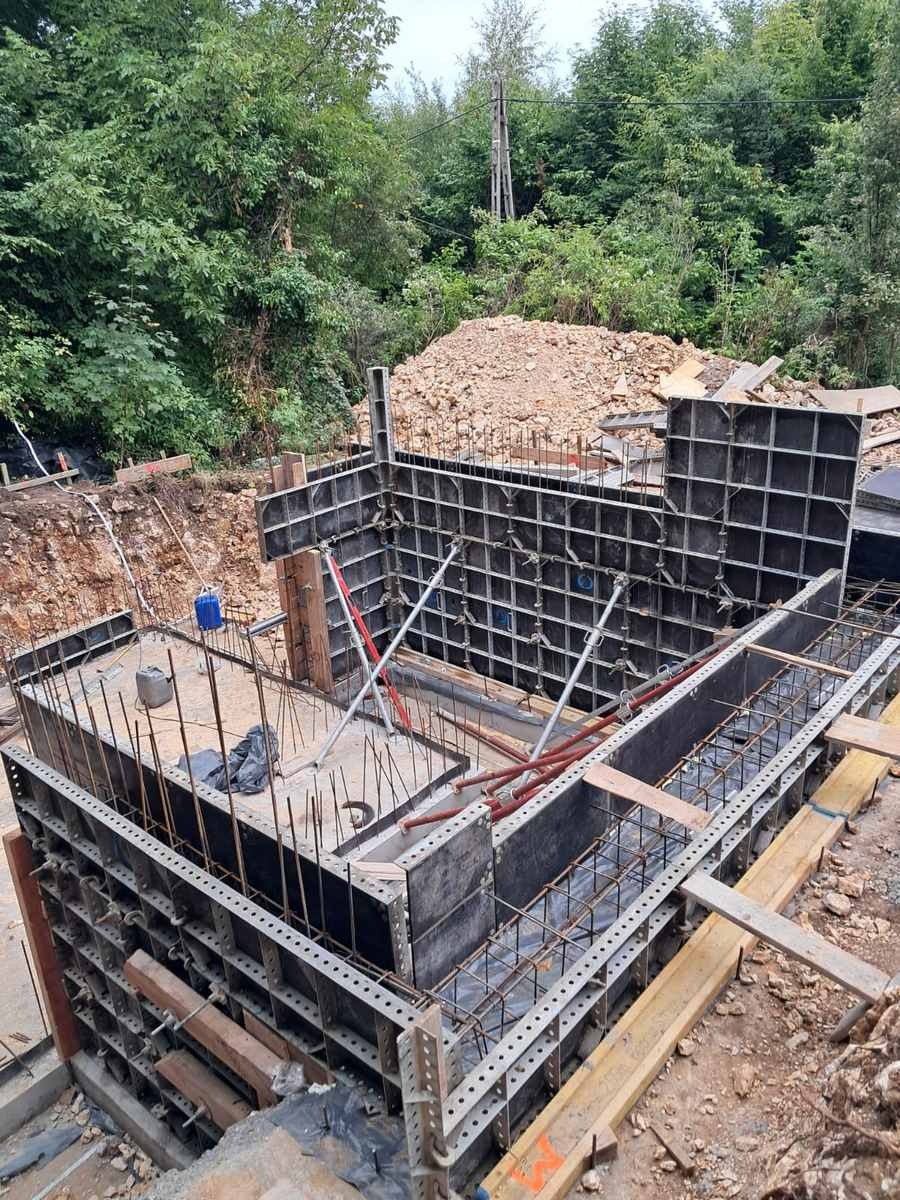 Szare szalunki fundamentów z widocznym zbrojeniem na tle zieleni i ziemi. Ujęcie z góry ukazuje detale konstrukcji i otoczenie placu budowy.