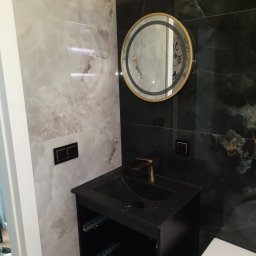 Michał Waszak - Wykończona łazienka z umywalką w czarnej szafce, okrągłym lustrem z podświetleniem i marmurowymi płytkami w odcieniach szarości i czerni. Nowoczesny design.