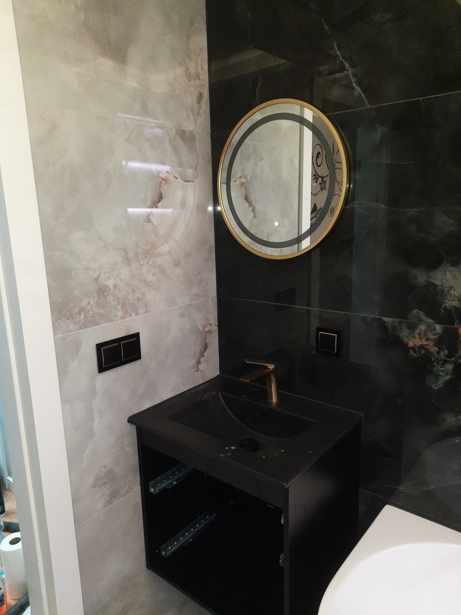 Wykończona łazienka z umywalką w czarnej szafce, okrągłym lustrem z podświetleniem i marmurowymi płytkami w odcieniach szarości i czerni. Nowoczesny design.