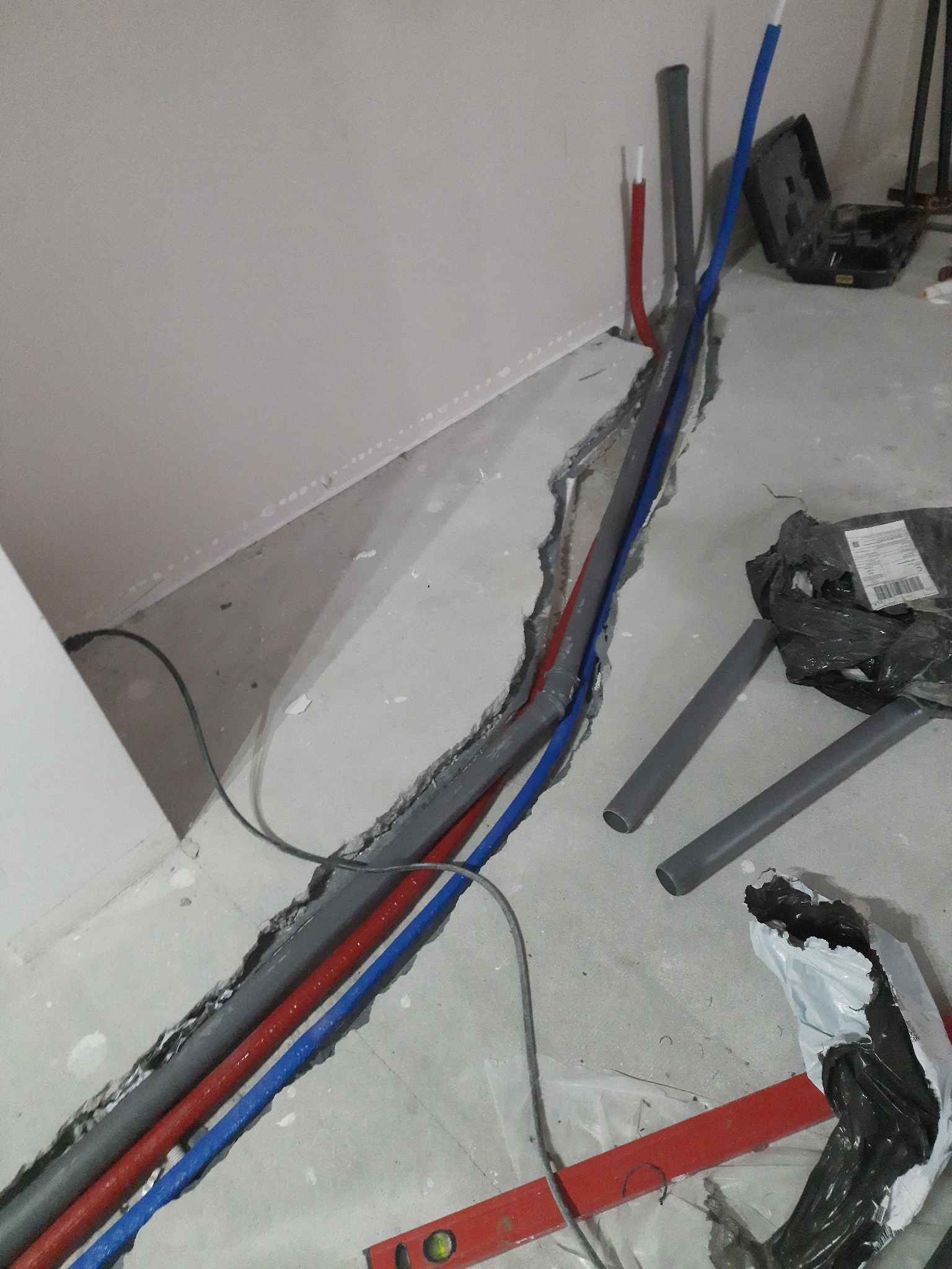 Instalacja wodna i kanalizacyjna w bruździe podłogowej, rury w kolorach niebieskim, czerwonym i szarym, poziomica, narzędzia w tle, surowy stan wykończenia.