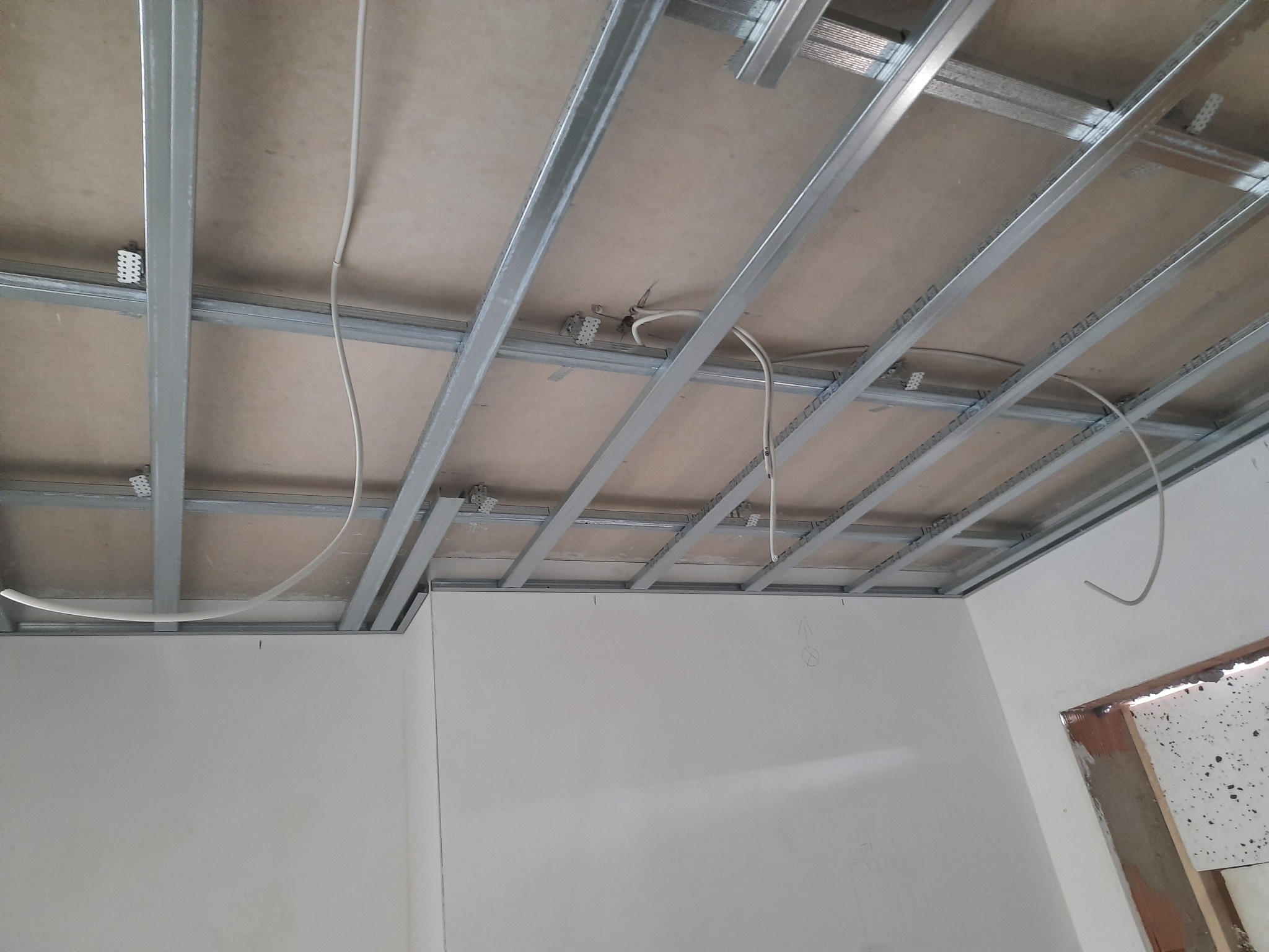 Sufit podwieszany w trakcie montażu: metalowa konstrukcja z widocznymi przewodami elektrycznymi, częściowo wykończona płyta karton-gips, surowe ściany w tle.