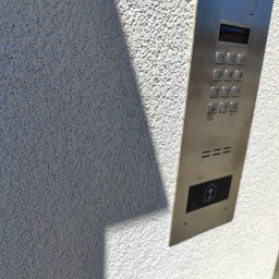 INOS Instalacje Mariusz Wojnik - Metalowy panel domofonu ACO z klawiaturą numeryczną i czytnikiem kart zbliżeniowych, zamontowany na jasnej, chropowatej ścianie budynku w słoneczny dzień.