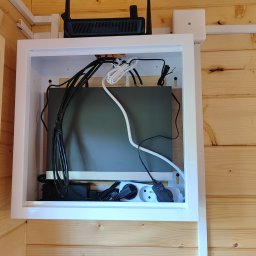 INOS Instalacje Mariusz Wojnik - Zamknięta, biała szafka z routerem, zasilaczami i kablami na drewnianej ścianie. Estetyczne ukrycie instalacji elektrycznej i sieciowej w domu.
