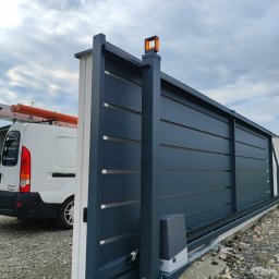 INOS Instalacje Mariusz Wojnik - Automatyczna brama przesuwna w kolorze grafitowym, z widocznym napędem i lampą ostrzegawczą. W tle biały bus z drabiną i rurami na dachu, sugerujący montaż lub serwis.