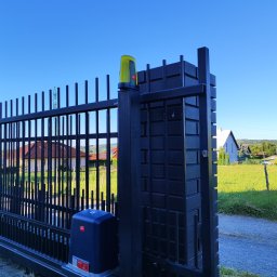 INOS Instalacje Mariusz Wojnik - Automatyczna brama wjazdowa z napędem BFT, widok z bliska na mechanizm i fragment ogrodzenia. W tle domy i zieleń, błękitne niebo. Nowoczesny design.