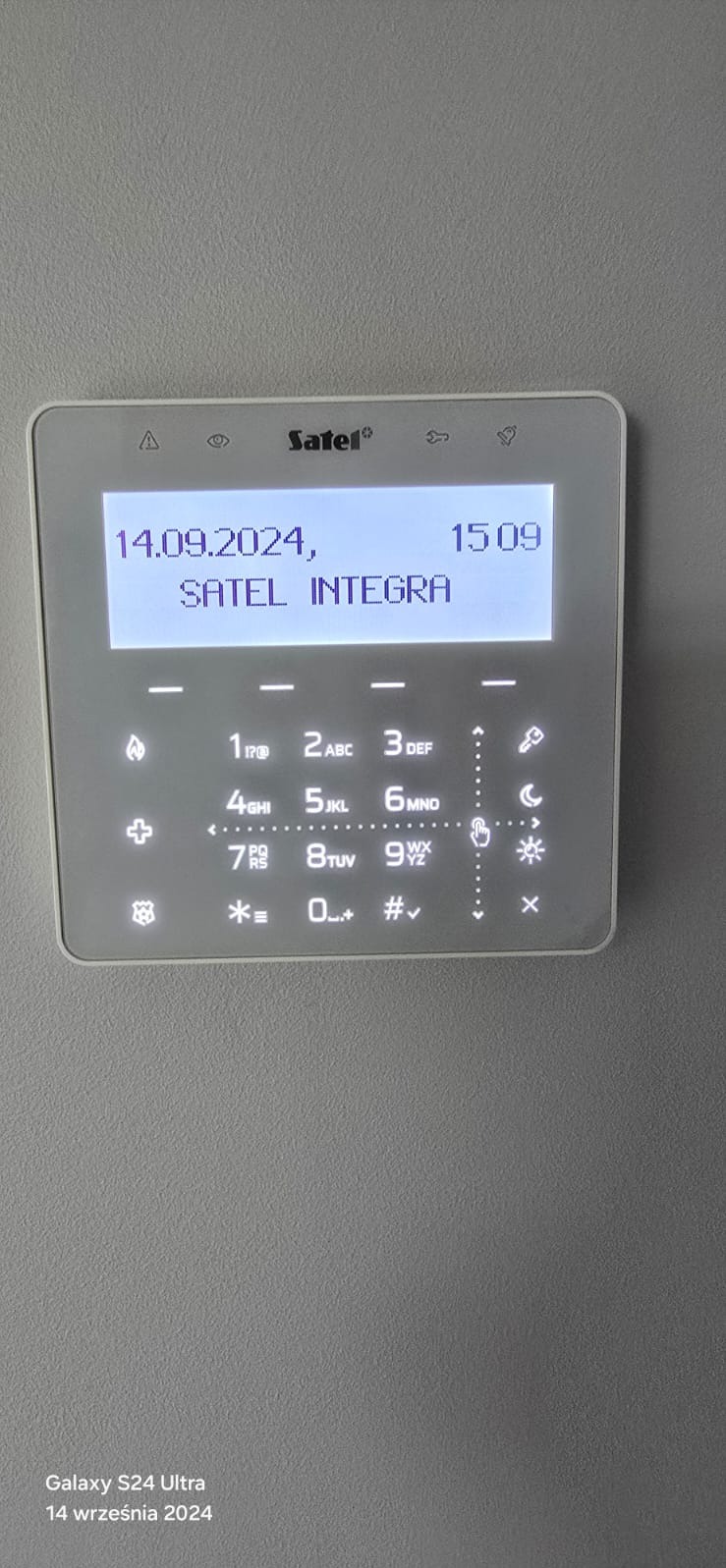 Biały panel alarmowy Satel Integra z podświetlonym ekranem LCD, wyświetlającym datę i godzinę. Widoczne przyciski numeryczne i funkcyjne. Szary, teksturowany mur w tle.