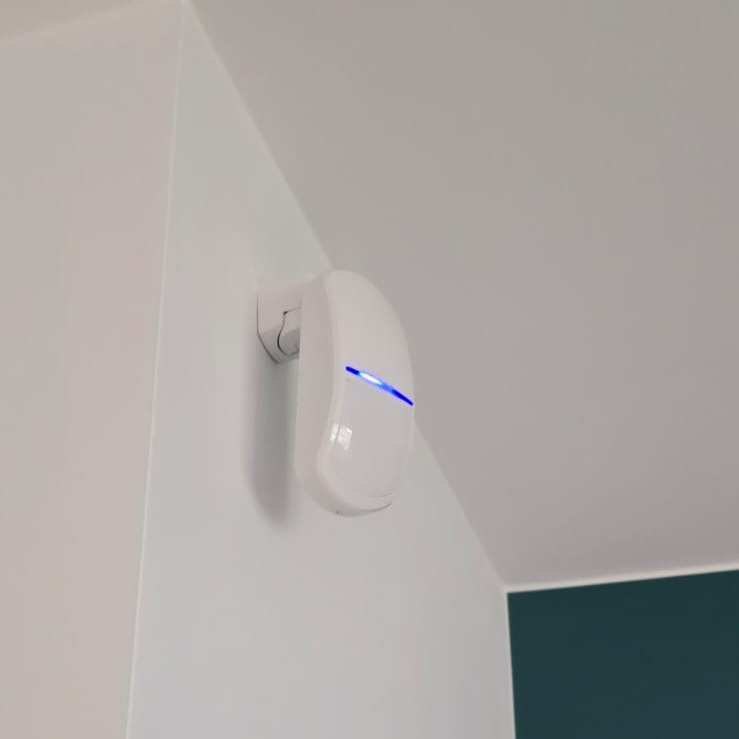 Biały czujnik alarmowy z niebieską diodą LED, zamontowany na białej ścianie w rogu pomieszczenia, widok z dołu.