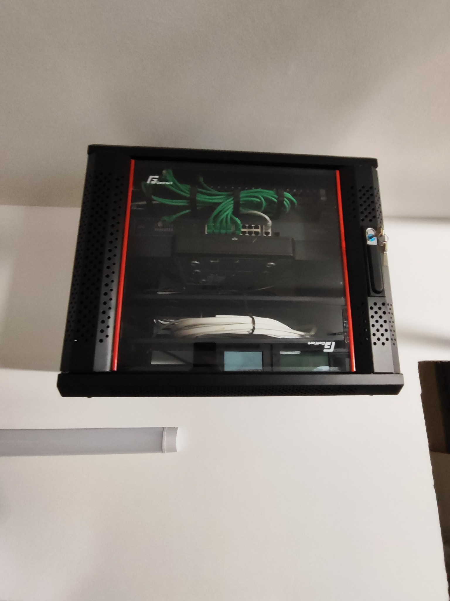 Zamknięta szafa rack wisząca na ścianie, z widocznymi kablami i urządzeniami sieciowymi wewnątrz. Oświetlenie sufitowe w tle. Widok z dołu.