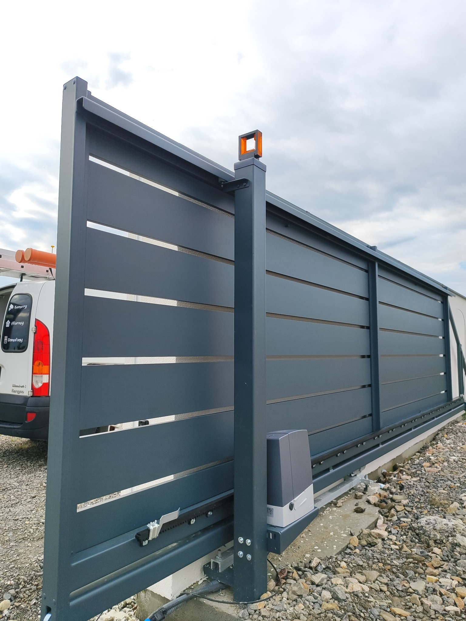 Nowoczesna, przesuwna brama wjazdowa z automatyką i lampą ostrzegawczą, widoczna z boku. Szara, pozioma konstrukcja z przerwami, na tle pochmurnego nieba.