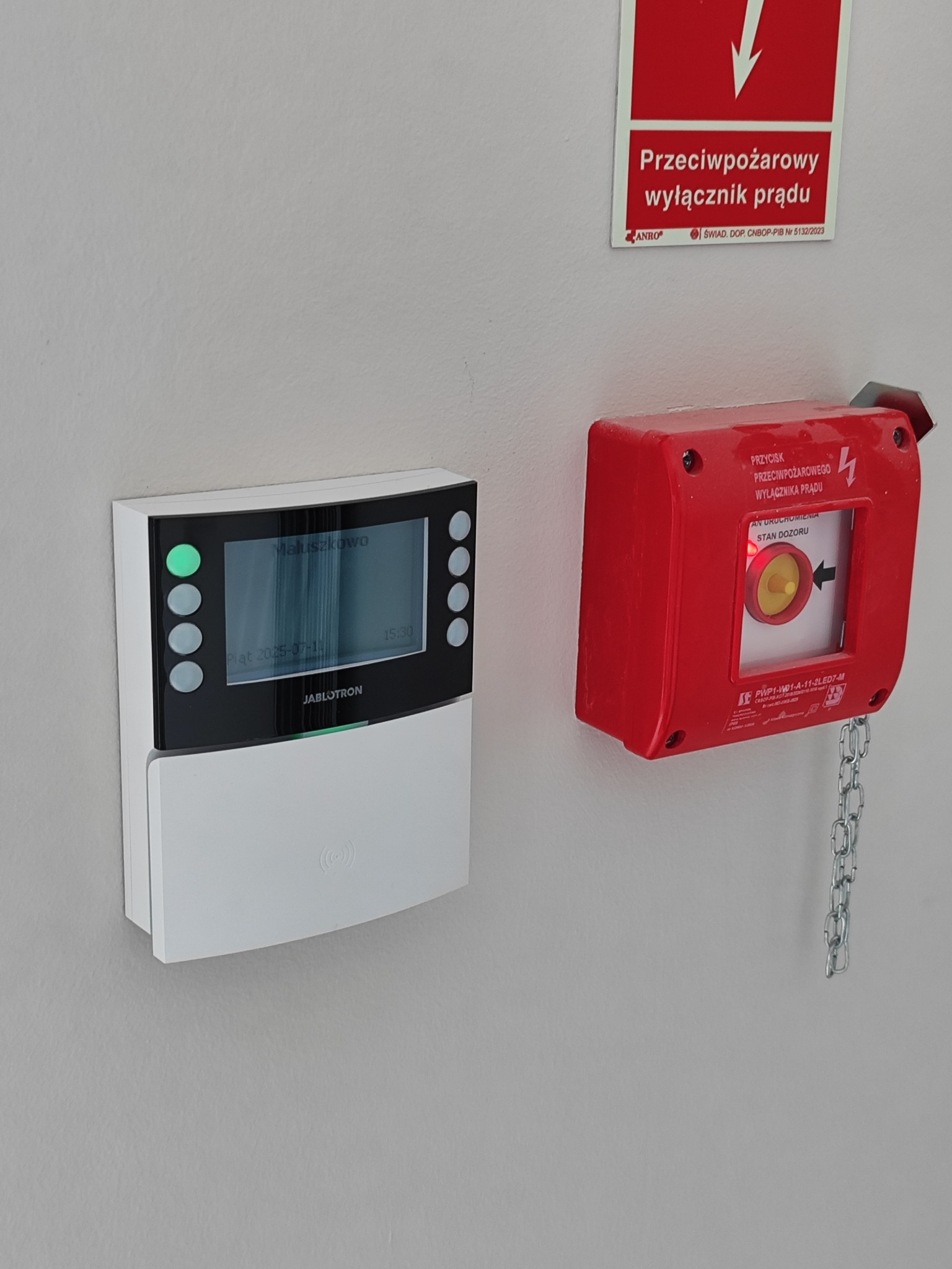 Panel alarmowy Jablotron z zielonymi diodami i czerwony przycisk przeciwpożarowy na ścianie. Widoczny wyświetlacz z datą i godziną. System alarmowy w budynku.