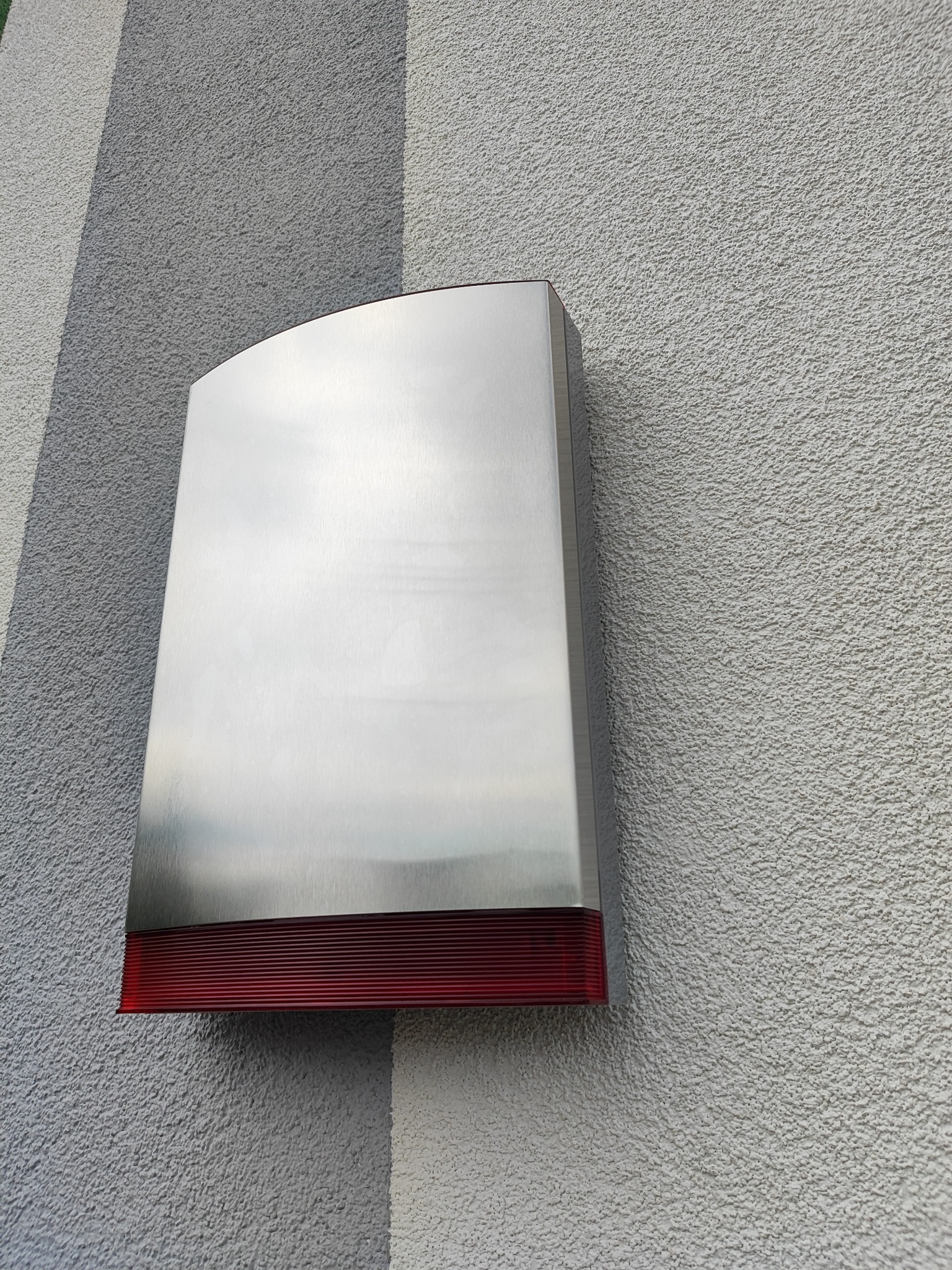 Zewnętrzna, metalowa obudowa alarmowa z czerwonym paskiem na dole, zamontowana na szarej, teksturowanej ścianie budynku. Minimalistyczny design, nowoczesne wykończenie.