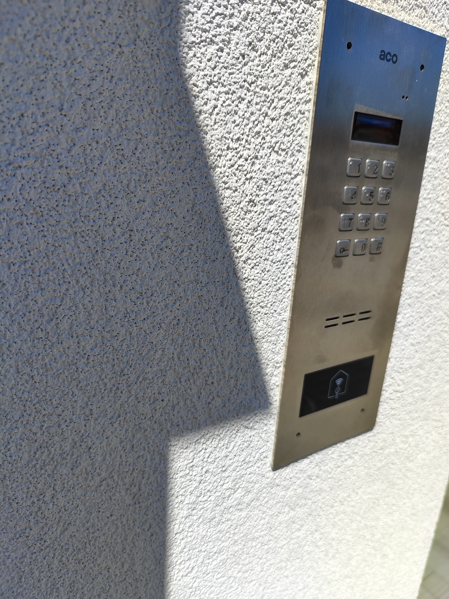 Metalowy panel domofonu ACO z klawiaturą numeryczną i czytnikiem kart zbliżeniowych, zamontowany na jasnej, chropowatej ścianie budynku w słoneczny dzień.