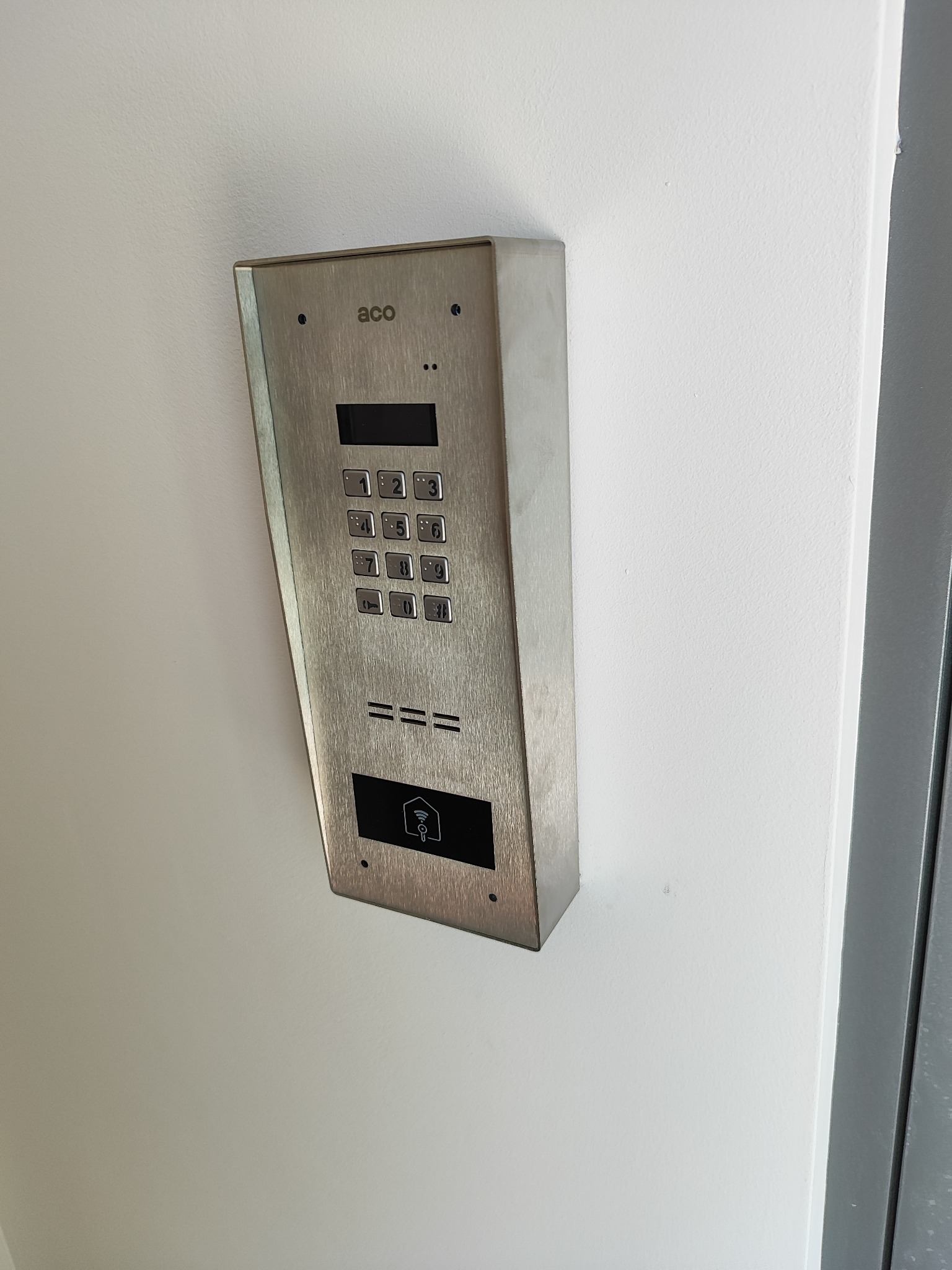 Zamontowany, metalowy panel domofonu ACO z klawiaturą numeryczną i czytnikiem kart zbliżeniowych na białej ścianie, obok szarej futryny drzwi.
