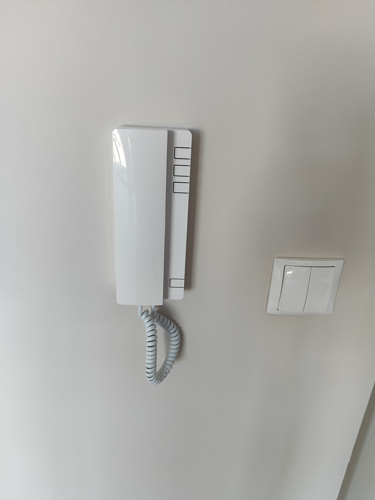 Biały domofon z kręconym kablem zamontowany na jasnej ścianie obok włącznika światła. Minimalistyczny design, czyste linie, nowoczesne wykończenie.