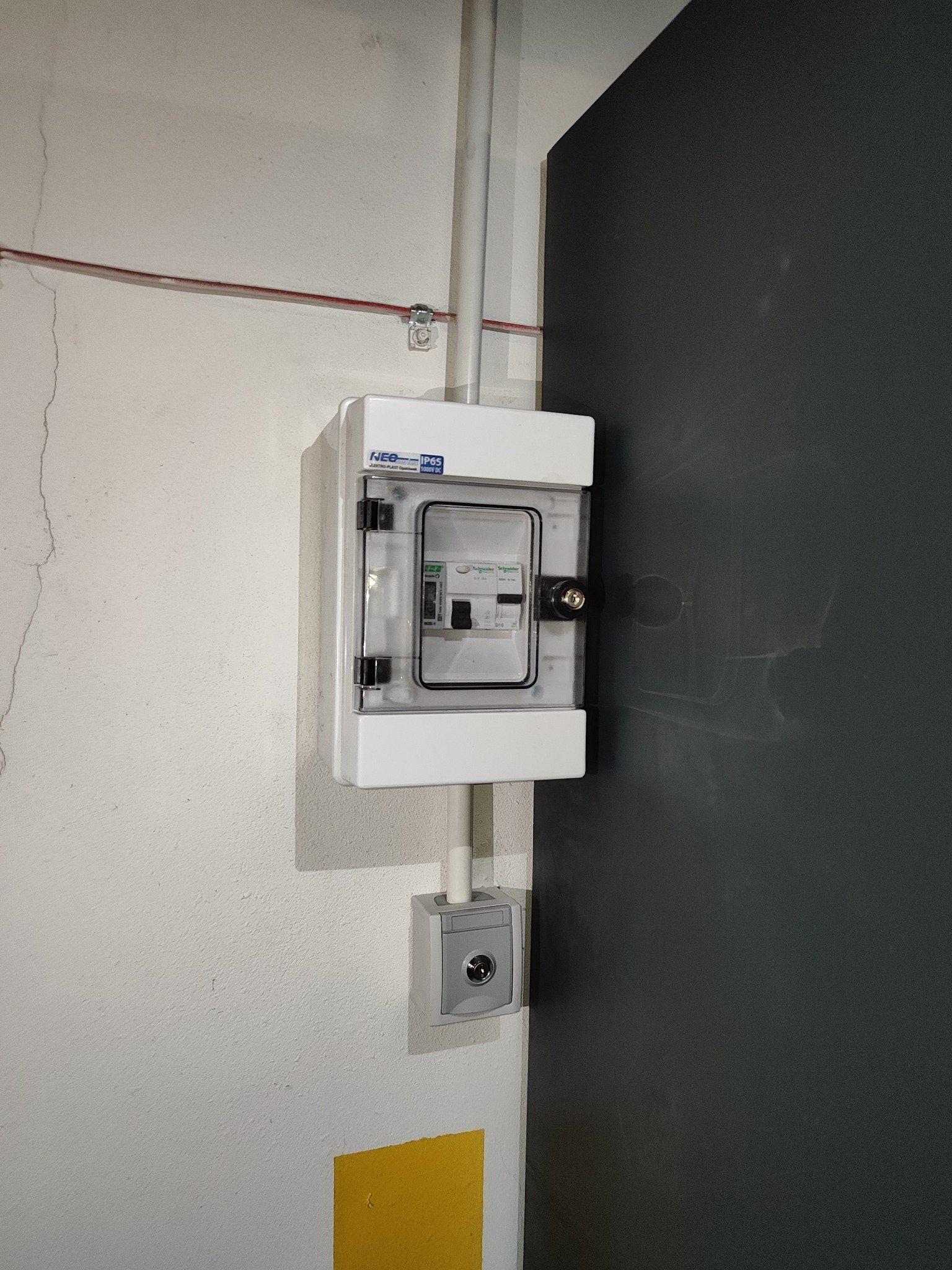 Zamknięta, biała skrzynka elektryczna z przezroczystymi drzwiczkami, zamontowana na białej ścianie obok ciemnoszarych drzwi. Widoczne przewody i dodatkowa puszka pod skrzynką.