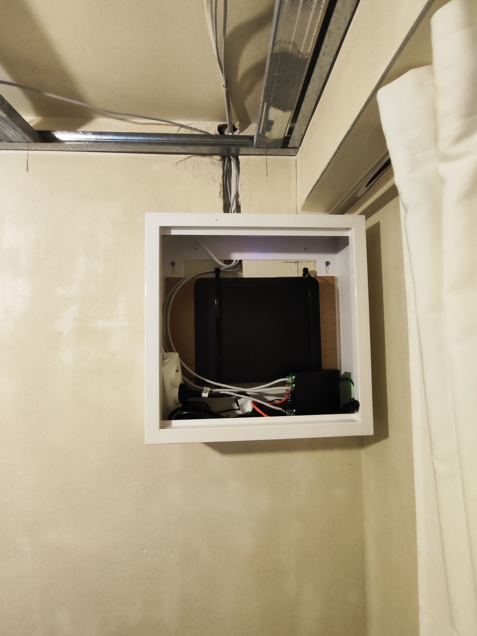 Zamknięta, biała szafka z czarnym urządzeniem i kablami na ścianie, widoczne elementy instalacji pod sufitem. Ujęcie z dołu, oświetlenie mieszane.