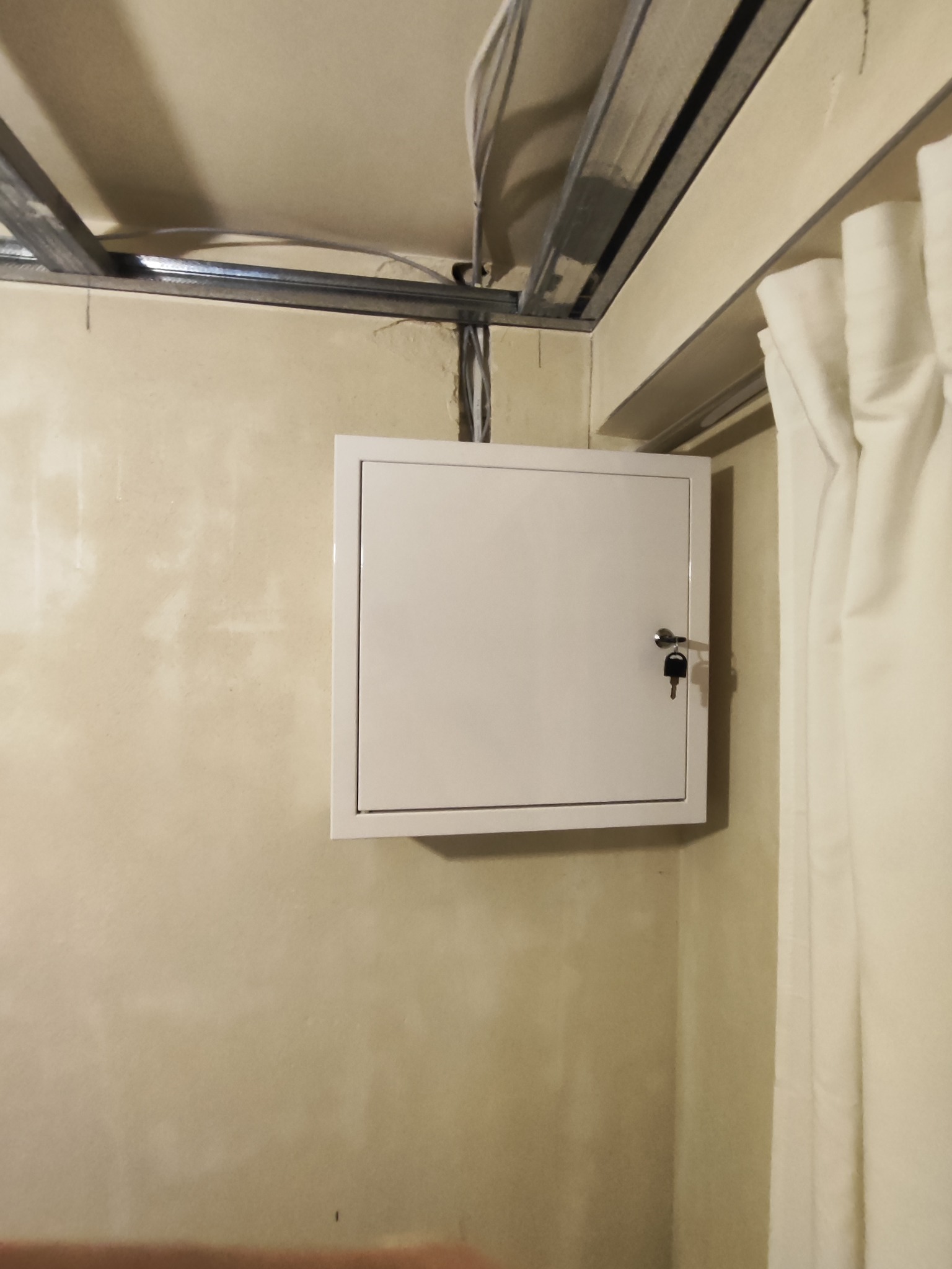 Zamknięta, biała skrzynka z kluczykiem, zamontowana na ścianie wewnątrz budynku. Widoczne przewody, sufit podwieszany i fragment zasłony. Ujęcie z dołu.