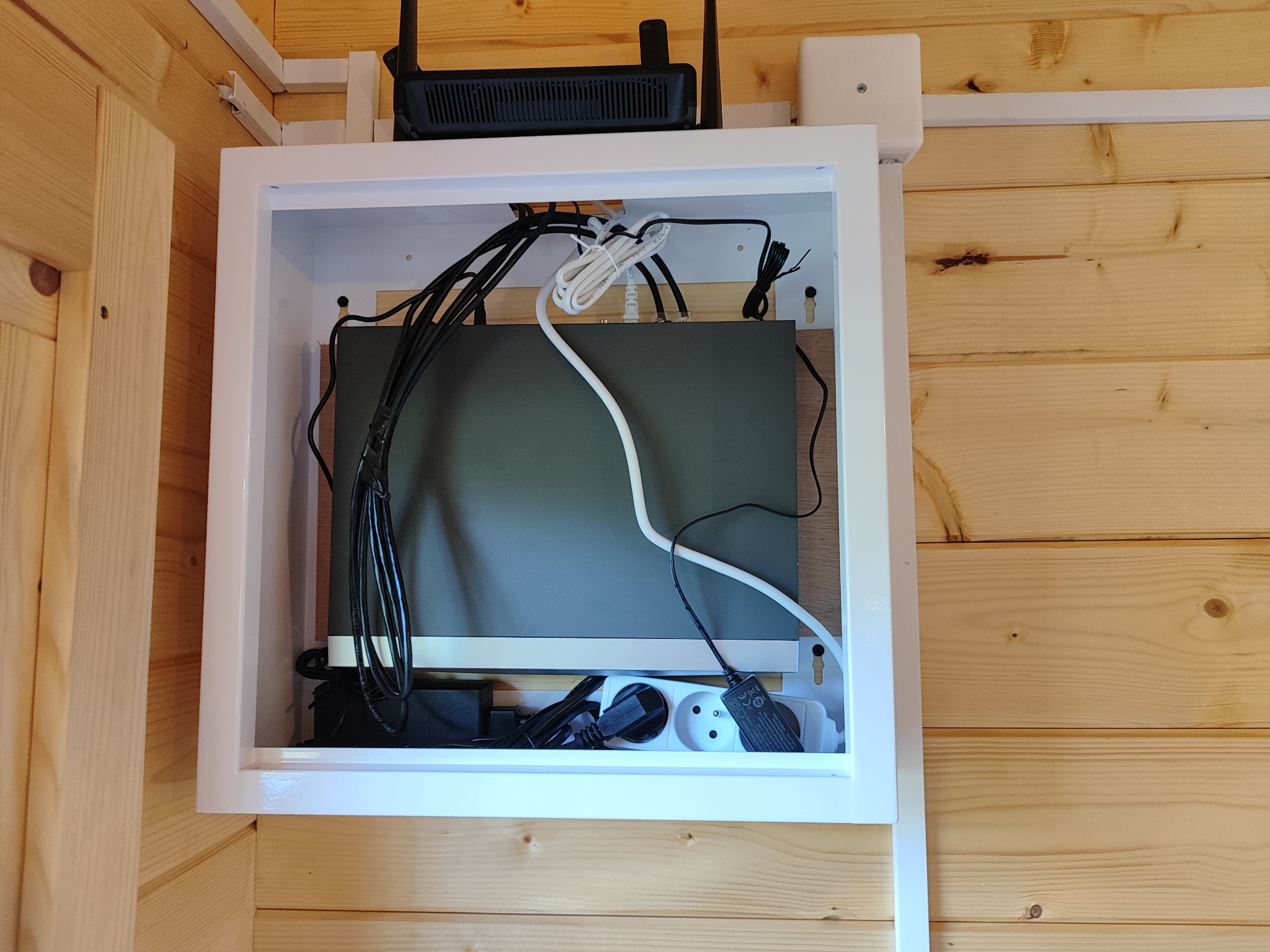 Zamknięta, biała szafka z routerem, zasilaczami i kablami na drewnianej ścianie. Estetyczne ukrycie instalacji elektrycznej i sieciowej w domu.
